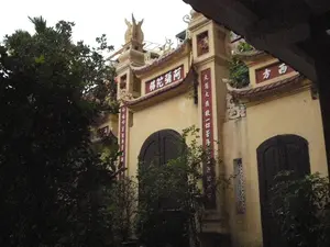 Bức ảnh chụp một phần của một ngôi chùa Phật giáo có kiến trúc cổ. Tòa nhà được sơn màu vàng kem, nổi bật với các chi tiết trang trí và chữ viết màu đỏ. Xung quanh có nhiều cây cối xanh tươi, tạo nên một khung cảnh tĩnh lặng và trang nghiêm.。**Thông tin chữ viết:**。Trên tòa nhà có các bức hoành phi và câu đối viết bằng chữ Hán:。- Bức hoành phi ở giữa: 「佛陀阿彌」 (Phật Đà A Di - A Di Đà Phật).。- Bức hoành phi bên phải: 「方西」 (Phương Tây - chỉ cõi Tây Phương Cực Lạc).。- Câu đối dọc bên trái: 「大慈大悲救一切苦海」 (Đại từ đại bi cứu nhất thiết khổ hải - Lòng từ bi vĩ đại cứu vớt tất cả khỏi bể khổ).。**Mô tả cảnh vật và hoạt động:**。- **Loại địa điểm:** Một ngôi chùa Phật giáo (Chùa).。- **Hoạt động:** Không có hoạt động nào của con người được ghi lại, khung cảnh rất tĩnh lặng.。- **Vật thể:** Tòa nhà có kiến trúc chùa chiền truyền thống với mái cong và các chi tiết trang trí trên đỉnh. Có các cửa gỗ lớn hình vòm đang đóng. Xung quanh là cây cối rậm rạp.。（日本語訳）。写真は、古い建築様式の仏教寺院の一部を写したものです。建物はクリームイエローに塗られ、赤い装飾や文字が際立っています。周囲には青々とした木々が多く、静かで荘厳な風景を醸し出しています。**文字情報：**。建物には漢字で書かれた扁額と対聯があります。- 中央の扁額：「佛陀阿彌」（Phật Đà A Di - 阿弥陀仏）。- 右側の扁額：「方西」（Phương Tây - 西方極楽浄土を指す）。- 左側の縦の対聯：「大慈大悲救一切苦海」（Đại từ đại bi cứu nhất thiết khổ hải - 大いなる慈悲は一切の苦海を救う）。**風景と活動の描写：**。- **場所の種類：** 仏教寺院（Chùa）。- **活動：** 人の活動は見られず、風景は非常に静かです。- **物体：** 建物は、湾曲した屋根と頂上の装飾を持つ伝統的な寺院建築です。大きなアーチ型の木製の扉は閉まっています。周囲は木々が鬱蒼と茂っています。