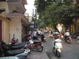 Đây là cảnh một con phố đông đúc ở Hà Nội vào buổi chiều tối. Lòng đường tấp nập người qua lại, chủ yếu bằng xe máy. Ở phía trước, một người phụ nữ đội mũ bảo hiểm màu xanh đang lái một chiếc xe tay ga màu trắng. Nhiều xe máy khác cũng đang lưu thông trên đường. Dọc vỉa hè bên trái, rất nhiều xe máy đang được dựng đỗ. Một nhóm người đang đứng tụ tập trên vỉa hè. Hai bên đường là những ngôi nhà san sát và có nhiều cây xanh lớn. Trên bức tường của ngôi nhà màu kem bên trái có một vài chữ viết màu đỏ nhưng không thể đọc được.。（日本語訳）。これは夕方のハノイの混雑した通りの光景です。道路は主にバイクで人々がひっきりなしに行き交っています。前方では、青いヘルメットをかぶった女性が白いスクーターを運転しています。他の多くのバイクも道路を走行しています。左側の歩道沿いには、たくさんのバイクが駐車されています。歩道では、あるグループの人々が立って集まっています。道路の両側には家々が密集しており、大きな緑の木々がたくさんあります。左側のクリーム色の家の壁には、いくつかの赤い文字が書かれていますが、読むことはできません。