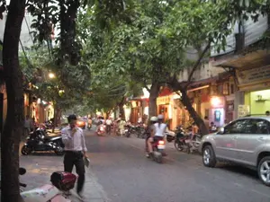 Bối cảnh là một con phố ở Hà Nội vào lúc chạng vạng. Ánh sáng từ đèn đường và các cửa hiệu đã bắt đầu bật lên. Hai bên đường có nhiều cây xanh lớn, tạo nên một không gian rợp bóng mát. Các tòa nhà san sát nhau, tầng trệt được sử dụng làm cửa hàng kinh doanh. Hoạt động giao thông trên đường khá nhộn nhịp với nhiều xe máy đang di chuyển. Một chiếc ô tô màu bạc đang đỗ ở lề đường bên phải. Ở phía trước, một người đàn ông mặc áo sơ mi kẻ sọc đang đi bộ trên vỉa hè và dường như đang nói chuyện điện thoại. Có nhiều người khác đang đi xe máy, một số người đội mũ bảo hiểm. Nhiều xe máy khác được dựng trên vỉa hè trước các cửa hàng. Có các biển hiệu của cửa hàng nhưng chữ viết bị mờ và không thể đọc rõ.。（日本語訳）。背景は、夕暮れ時のハノイのある通りです。街灯や店の明かりが灯り始めています。道の両側には大きな緑の木々が多く、日陰の空間を作り出しています。建物が密集しており、1階は店舗として使われています。道路の交通はかなり賑わっており、多くのバイクが移動しています。銀色の車が道路の右側に駐車しています。前景には、ストライプのシャツを着た男性が歩道を歩いており、電話で話しているようです。他にも多くの人がバイクに乗っており、何人かはヘルメットをかぶっています。他の多くのバイクが店の前の歩道に停められています。店の看板がありますが、文字はぼやけていてはっきりと読むことはできません。