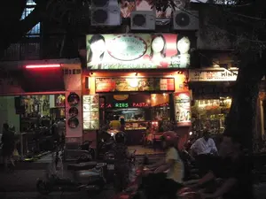 Bức ảnh chụp cảnh một con phố vào ban đêm, có nhiều cửa hàng đang hoạt động.。【Thông tin chữ viết】。*   Biển hiệu chính của một nhà hàng: "PEPPERONI'S", "PIZZA & CAFE", "1992".。*   Biển hiệu đèn neon: "PIZZA RIBS PASTA".。*   Biển hiệu quảng cáo: "FREE DELIVERY。".。*   Biển hiệu thực đơn phía trên quầy có các dòng chữ tiếng Việt, một số có thể đọc được như "MỲ Ý & SALAD...", "...KIỂU TUỲ THÍCH" và giá tiền "48.000".。*   Cửa hàng bên cạnh ở địa chỉ số 31 có biển hiệu "MINH ART ANTIQUE".。【Mô tả cảnh vật và hoạt động】。*   **Địa điểm:** Đây là một con phố thương mại sầm uất vào ban đêm. Trọng tâm của bức ảnh là nhà hàng pizza "Pepperoni's" và một cửa hàng đồ cổ "Minh Art Antique" ở bên cạnh. Phía trên các cửa hàng có gắn nhiều cục nóng điều hòa không khí.。*   **Hoạt động:** Các cửa hàng đang mở cửa kinh doanh. Bên trong nhà hàng pizza, có thể thấy nhân viên mặc đồng phục màu vàng đang làm việc sau quầy. Bên ngoài, đường phố tấp nập người qua lại bằng xe máy. Một số người đang ngồi trên xe máy, trong khi những người khác đang di chuyển, tạo ra vệt mờ trong ảnh. Nhiều xe máy cũng đang được đỗ ở lề đường trước các cửa hàng.。*   **Vật thể:** Các biển hiệu quảng cáo được chiếu sáng rực rỡ, xe máy (phương tiện di chuyển và đỗ), cây cối ven đường, và các tòa nhà san sát nhau. Không khí chung cho thấy một đêm nhộn nhịp của cuộc sống thành thị.。（日本語訳）。夜の通りを撮影した写真で、多くの店が営業している。【文字情報】。*   あるレストランのメイン看板：「PEPPERONI'S」、「PIZZA & CAFE」、「1992」。*   ネオンサイン：「PIZZA RIBS PASTA」。*   広告看板：「FREE DELIVERY。」。*   カウンターの上にあるメニュー看板にはベトナム語の文字があり、その一部は「MỲ Ý & SALAD...」（スパゲッティ＆サラダ...）、「...KIỂU TUỲ THÍCH」（...お好みのスタイルで）や価格「48.000」などが読み取れる。*   住所31番地にある隣の店には「MINH ART ANTIQUE」という看板がある。【風景と活動の描写】。*   **場所：** これは夜の賑やかな商店街である。写真の中心はピザレストラン「Pepperoni's」と隣にある骨董品店「Minh Art Antique」である。*   **活動：** 各店は営業中である。外では、通りはバイクで行き交う人々で混雑している。バイクに座っている人もいれば、移動中の人もおり、写真にブレた軌跡を残している。多くのバイクが店の前の歩道脇に駐車されている。*   **物体：** 明るく照らされた広告看板、バイク（移動手段および駐車中のもの）、街路樹、そして密集した建物。全体的な雰囲気は、都市生活の賑やかな夜を示している。