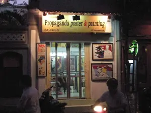 Bối cảnh là một con phố vào ban đêm, có thể là ở Hà Nội. Điểm nổi bật là mặt tiền của một cửa hàng nghệ thuật.。**Chữ viết được ghi lại:**。Trên biển hiệu được chiếu sáng của cửa hàng có ghi:。- Tên cửa hàng: "Propaganda poster & painting"。- Địa chỉ: "25A Lý Quốc Sư"。- Số điện thoại: "ĐT: 0912662779"。- Phía trên có một tên riêng khác nhưng khó đọc, có thể là "Lưu Manh".。- Trên một bức tranh có dòng chữ nhưng không thể đọc rõ.。**Mô tả cảnh vật và hoạt động:**。- **Loại địa điểm:** Đây là một cửa hàng nghệ thuật hoặc phòng trưng bày nằm trên một con phố (phố Lý Quốc Sư) ở Hà Nội. Cửa hàng chuyên bán các tác phẩm tranh và áp phích theo phong cách tuyên truyền.。- **Hoạt động cụ thể:** Cửa hàng đang mở cửa kinh doanh vào buổi tối. Bên trong cửa hàng có người đang xem hoặc làm việc. Bên ngoài, một người đang điều khiển xe máy đi trên đường với đèn pha bật sáng. Một người khác đang đứng trên vỉa hè gần một chiếc xe máy đang đỗ. Đây là một cảnh sinh hoạt đường phố về đêm.。- **Vật thể trong ảnh:**。- **Biển hiệu cửa hàng:** Một biển hiệu lớn, hình chữ nhật, được chiếu sáng, ghi rõ tên, địa chỉ và số điện thoại của cửa hàng.。- **Tranh và áp phích:** Nhiều bức tranh và áp phích theo phong cách tuyên truyền được đóng khung cẩn thận và treo trên tường bên ngoài cũng như trưng bày bên trong cửa hàng. Nổi bật là hai bức tranh lớn bên phải cửa ra vào; bức trên có vẻ là chân dung của Chủ tịch Hồ Chí Minh, bức dưới là hình một người phụ nữ trên nền cờ.。- **Xe máy:** Có ít nhất hai chiếc xe máy trong ảnh, một phương tiện giao thông phổ biến ở Việt Nam.。（日本語訳）。背景は夜の街路で、おそらくハノイでしょう。際立っているのは、あるアートショップの正面です。**記録された文字：**。店の照らされた看板には、次のように書かれています：。- 店名：「Propaganda poster & painting」。- 住所：「25A Lý Quốc Sư」。- 電話番号：「ĐT: 0912662779」。- 上部には別の固有名詞がありますが、読みにくく、おそらく「Lưu Manh」でしょう。- ある絵には文字がありますが、はっきりと読むことはできません。**風景と活動の説明：**。- **場所の種類：** これはハノイのある通り（Lý Quốc Sư通り）にあるアートショップまたはギャラリーです。この店はプロパガンダ様式の絵画やポスターの販売を専門としています。- **具体的な活動：** 店は夜に営業しています。店内には見ている人や作業している人がいます。外では、一人がヘッドライトを点灯させてバイクを運転しています。もう一人は、駐車しているバイクの近くの歩道に立っています。これは夜の街の日常風景です。- **写真の中の物体：**。- **店の看板：** 大きな長方形の照らされた看板で、店の名前、住所、電話番号がはっきりと記されています。- **絵画とポスター：** 多くのプロパガンダ様式の絵画やポスターが丁寧に額装され、外壁に掛けられたり、店内に展示されたりしています。入り口の右側にある2枚の大きな絵が目立ちます。上の絵はホー・チ・ミン主席の肖像画のようで、下の絵は旗を背景にした女性の姿です。- **バイク：** 写真には少なくとも2台のバイクが写っており、これはベトナムで一般的な交通手段です。