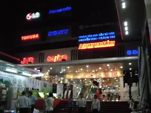 Bức ảnh chụp cảnh ban đêm phía trước một trung tâm mua sắm điện máy lớn, có tên là Nguyễn Kim. Tòa nhà được chiếu sáng rực rỡ bởi nhiều biển hiệu đèn neon.。Các dòng chữ có thể đọc được bao gồm:。- Tên các thương hiệu điện tử nổi tiếng trên mặt tiền tòa nhà: LG, Panasonic, TOSHIBA, SONY, JVC, SANYO.。- Biển hiệu chính của trung tâm: "TRUNG TÂM MUA SẮM SÀI GÒN NGUYỄN KIM - TRÀNG THI", với tên "Nguyễn Kim" được làm nổi bật bằng đèn màu đỏ.。- Một biểu ngữ quảng cáo ở bên trái ghi: "Hệ Khử Mùi & Diệt Khuẩn HYBRID PLASMA".。- Bên trong cửa hàng có một biển hiệu ghi "TOSHIBA".。Đây là một trung tâm thương mại nhiều tầng. Lối vào chính có cầu thang bộ trải thảm đỏ. Bên trong có thể nhìn thấy thang cuốn, các quầy hàng trưng bày sản phẩm điện tử như tủ lạnh, và nhiều người đang đi lại. Bên ngoài và bên trong cửa hàng có một số người, có vẻ là khách hàng và nhân viên mặc đồng phục, đang đứng hoặc di chuyển, cho thấy hoạt động mua sắm và tham quan đang diễn ra. Phía bên phải lối vào có đặt một vài bức tượng trang trí màu trắng.。（日本語訳）。グエンキムという名前の大きな家電ショッピングセンターの前で撮影された夜景の写真です。建物は多くのネオンサインによって煌々と照らされています。読み取れる文字には以下が含まれます：。- 建物のファサードにある有名な電子機器ブランド名：LG、Panasonic、TOSHIBA、SONY、JVC、SANYO。- センターのメイン看板：「TRUNG TÂM MUA SẮM SÀI GÒN NGUYỄN KIM - TRÀNG THI」、その中で「Nguyễn Kim」の名前は赤い光で際立たされています。- 左側の広告バナーには「Hệ Khử Mùi & Diệt Khuẩn HYBRID PLASMA」と書かれています。- 店内には「TOSHIBA」と書かれた看板があります。これは多層階の商業施設です。メインの入り口には赤いカーペットが敷かれた階段があります。内部では、エスカレーター、冷蔵庫などの電子製品を展示する売り場、そして多くの人々が行き来しているのが見えます。店の外と中には、客や制服を着た従業員と見られる何人かの人々が立っていたり移動したりしており、買い物や見学の活動が行われていることを示しています。入り口の右側には、いくつかの白い装飾的な像が置かれています。
