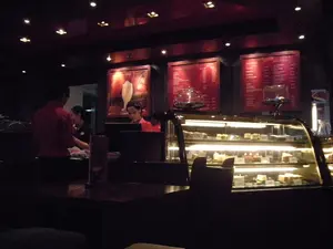 Bối cảnh là bên trong một quán cà phê có không gian tối và ấm cúng. Các nhân viên đang làm việc phía sau quầy phục vụ. Một người đàn ông mặc áo sơ mi đỏ đang đứng quay lưng lại, và hai nhân viên nữ đang ở gần đó; một người đang thao tác trên máy tính có vẻ là máy tính tiền. Phía trước quầy là một tủ kính lớn được chiếu sáng, trưng bày nhiều loại bánh ngọt khác nhau trên nhiều tầng. Phía trên và phía sau quầy, có ba bảng thực đơn lớn có đèn nền màu đỏ. Chữ trên các bảng thực đơn này không thể đọc được, nhưng một bảng có hình ảnh một ly đồ uống đá xay. Ánh sáng trong quán khá yếu, chủ yếu đến từ các đèn âm trần và ánh sáng từ tủ trưng bày bánh, tạo nên một không khí sang trọng.。（日本語訳）。背景は、暗くて居心地の良いカフェの店内です。スタッフがカウンターの後ろで働いています。赤いシャツを着た男性が背を向けて立っており、その近くに2人の女性スタッフがいます。1人はレジと思われるコンピューターを操作しています。カウンターの前には、照明付きの大きなガラスケースがあり、様々な種類のケーキが何段にもわたって陳列されています。カウンターの上と後ろには、赤いバックライトが付いた3つの大きなメニューボードがあります。これらのメニューボードの文字は読めませんが、1つのボードにはフラッペドリンクのグラスの絵があります。店内の照明はかなり暗く、主に埋め込み式の天井照明とケーキのショーケースからの光で、高級感のある雰囲気を醸し出しています。