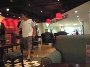 Bối cảnh là bên trong một không gian có vẻ như là một quán cà phê hoặc nhà hàng ở Hà Nội, với không khí ấm cúng và ánh sáng hơi tối. Không gian được trang bị nhiều loại bàn ghế, bao gồm ghế sofa lớn bọc nỉ ở phía trước, bàn ghế gỗ và ghế bành bọc da ở phía sau. Trần nhà có đèn âm trần và treo những chiếc đèn lồng hình trụ màu đỏ. Một người đàn ông mặc áo phông trắng và quần short đang đi bộ trong quán. Có những người khác cũng có mặt, bao gồm một người đàn ông đứng sau anh ta và một người phụ nữ ở phía bên trái. Sàn nhà có một khu vực được lát gạch hoa văn ca rô. Trên các bức tường có chữ viết trang trí, một bức tường màu đỏ ở hậu cảnh và một bức tường trắng ở phía xa, nhưng các ký tự đều bị mờ và không thể đọc rõ.。（日本語訳）。背景はハノイにあるカフェかレストランのような空間の中、暖かく、やや薄暗い雰囲気です。空間には様々な種類のテーブルと椅子が備え付けられており、手前には大きなフェルト張りのソファ、奥には木製のテーブルと椅子、革張りのアームチェアが含まれます。天井にはダウンライトがあり、赤い円筒形の提灯が吊るされています。白いTシャツとショートパンツを履いた男性が店の中を歩いています。他にも人がおり、彼の後ろに立つ男性と、左側にいる女性が含まれます。床には市松模様のタイルが敷かれたエリアがあります。壁には装飾的な文字が書かれており、背景には赤い壁、遠くには白い壁がありますが、文字はすべてぼやけていて、はっきりと読むことはできません。