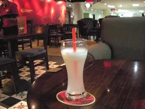 Bối cảnh là bên trong một quán cà phê hoặc nhà hàng có không gian hiện đại. Ở cận cảnh, trên một chiếc bàn gỗ sẫm màu, có một ly thủy tinh cao chứa đồ uống màu trắng đục, có vẻ như là một loại sinh tố hoặc nước dừa, với một ống hút màu đỏ. Ly nước được đặt trên một chiếc đế lót ly màu đỏ. Sàn nhà được lát gạch hoa văn ca-rô đen trắng xen kẽ với gạch hoa văn màu đỏ và trắng. Ở phía sau, có thể thấy nhiều bàn ghế hơn, bao gồm ghế đẩu bằng gỗ và ghế bành, sofa bọc nệm. Một người dường như là nhân viên, mặc áo sơ mi màu đỏ, đang đứng ở hậu cảnh. Phía sau người nhân viên là một bức tường màu đỏ được chiếu sáng, trên đó có một logo hình tròn nhưng không thể đọc rõ. Không gian chung có ánh sáng dịu, tạo cảm giác ấm cúng và thư giãn.。（日本語訳）。背景はモダンな空間のカフェまたはレストランの中です。手前では、濃い色の木製テーブルの上に、スムージーかココナッツウォーターと思われる不透明な白い飲み物が入った背の高いグラスがあり、赤いストローが添えられています。グラスは赤いコースターの上に置かれています。床は、白黒の市松模様のタイルと、赤と白の模様のタイルが交互に敷き詰められています。奥には、木製のスツールやアームチェア、布張りのソファなど、さらに多くのテーブルと椅子が見えます。店員と思われる人物が、赤いシャツを着て、奥に立っています。空間全体は柔らかな光で、居心地の良いリラックスした雰囲気を醸し出しています。