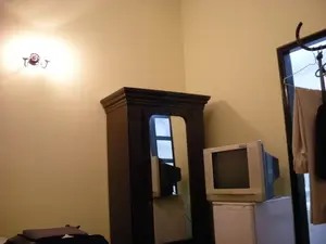 Bức ảnh chụp nội thất của một căn phòng, có thể là phòng khách sạn. Tường phòng được sơn màu kem. Trong phòng có một chiếc tủ quần áo bằng gỗ sẫm màu với một chiếc gương ở mặt trước. Bên cạnh tủ là một chiếc tivi CRT kiểu cũ được đặt trên một chiếc tủ lạnh mini màu trắng. Trên góc phải của tivi có một tem dán nhỏ nhưng không đọc được chữ. Trên tường bên trái, một chiếc đèn tường đang bật, tỏa ra ánh sáng vàng ấm. Ở phía bên phải của bức ảnh là một ô cửa, nơi có treo quần áo trên móc. Ở góc dưới bên trái, có thể thấy một phần của hành lý.。（日本語訳）。ある部屋の内部を撮影した写真で、ホテルの部屋かもしれません。部屋の壁はクリーム色に塗られています。部屋には、前面に鏡が付いた濃い色の木製クローゼットが1つあります。クローゼットの隣には、白いミニ冷蔵庫の上に置かれた旧式のブラウン管テレビがあります。テレビの右上に小さなシールが貼られていますが、文字は読めません。左側の壁では、壁掛けランプが点灯しており、暖かい黄色の光を放っています。写真の右側には戸口があり、そこにはハンガーにかけられた服があります。左下隅には、荷物の一部が見えます。