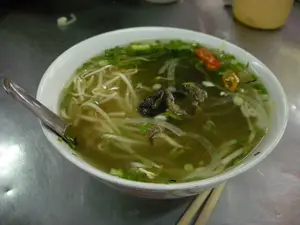 Đây là hình ảnh cận cảnh một tô phở bò, một món ăn đặc trưng của Việt Nam. Món ăn được đặt trên một chiếc bàn inox, cho thấy bối cảnh có thể là một quán ăn bình dân hoặc quán phở. Trong tô sứ màu trắng chứa đầy nước dùng trong, có thể thấy rõ các thành phần như bánh phở, giá đỗ, những miếng thịt bò, hành lá thái nhỏ, hành tây thái mỏng và vài lát ớt đỏ tươi. Một chiếc thìa inox được đặt trong tô, và một đôi đũa được đặt bên cạnh, sẵn sàng để thực khách thưởng thức. Không có văn bản hay chữ viết nào trong hình ảnh.。（日本語訳）。これは牛肉のフォーのクローズアップ写真で、ベトナムの代表的な料理です。料理はステンレス製のテーブルの上に置かれており、庶民的な食堂かフォー専門店といった背景がうかがえます。白い磁器の丼は透明なスープで満たされており、フォーの麺、もやし、牛肉のスライス、刻みネギ、薄切り玉ねぎ、そして数切れの新鮮な赤唐辛子といった具材がはっきりと見えます。ステンレス製のスプーンが丼の中に、そして箸一膳がその横に置かれ、食事客が味わう準備が整っています。画像内にはテキストや文字はありません。