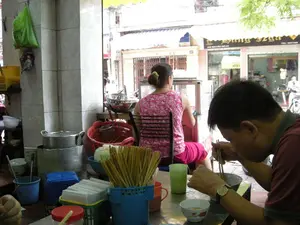 Chữ viết: Trên tấm biển hiệu của cửa hàng ở phía đối diện có ghi địa chỉ "41E MAI HẮC ĐẾ" và số điện thoại "TEL: 8227399". Tên cửa hàng có chữ "HÀNG SỰA" nhưng phần đầu không rõ.。Cảnh vật và hoạt động: Bức ảnh chụp bên trong một quán ăn ven đường ở Hà Nội. Một người đàn ông mặc áo đỏ đang ngồi ăn, dùng đũa gắp thức ăn từ một chiếc bát. Trên bàn có các vật dụng quen thuộc như hộp đựng đũa, giấy ăn, lọ gia vị, và một chiếc cốc nhựa màu xanh. Ở phía sau, một người phụ nữ mặc áo hoa đang ngồi quay lưng lại, gần khu vực nấu nướng với các nồi nhôm lớn, rổ nhựa và các dụng cụ nhà bếp khác. Khung cảnh nhìn ra đường phố, cho thấy một cửa hàng khác ở phía đối diện và một phần của cuộc sống đô thị thường nhật.。（日本語訳）。文字：向かいの店の看板には「41E MAI HẮC ĐẾ」という住所と「TEL: 8227399」という電話番号が書かれている。店名には「HÀNG SỰA」という文字があるが、最初の部分は不明瞭である。風景と活動：写真はハノイの道端にある食堂の内部を撮影したものである。赤いシャツを着た男性が座って食事をしており、箸を使って椀から食べ物を取っている。テーブルの上には、箸入れ、ティッシュ、調味料入れ、そして青いプラスチックのコップといったおなじみの物がある。後方では、花柄のシャツを着た女性が背を向けて座っており、大きなアルミ鍋、プラスチックのかご、その他の厨房用品がある調理場の近くにいる。