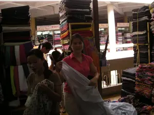 Bối cảnh là một khu chợ bán vải trong nhà, có vẻ đông đúc và nhộn nhịp. Hoạt động chính là mua bán vải. Một người phụ nữ mặc áo hồng (có thể là người bán hàng) đang tươi cười và giơ một tấm vải màu sáng cho khách xem. Một người phụ nữ khác mặc áo màu nâu (khách hàng) đang xem xét một mảnh vải có hoa văn. Xung quanh có nhiều người khác đang đi lại giữa các quầy hàng. Các vật thể nổi bật là vô số súc vải với đủ loại màu sắc, được xếp thành những chồng cao, có nơi gần chạm trần nhà. Ở phía sau, có thể thấy các biển hiệu của các quầy hàng, một trong số đó có ghi chữ 'QUẦY VẢI Hằng Thuỷ' và một biển hiệu khác có chữ 'NGỌC C...' nhưng bị che một phần. Các biển hiệu khác ở xa hơn thì không thể đọc rõ.。（日本語訳）。背景は屋内の生地市場で、混雑して活気があるようです。主な活動は生地の売買です。ピンクの服を着た女性（おそらく店員）が微笑みながら、明るい色の生地を客に見せています。別の茶色い服を着た女性（客）が、柄物の生地を吟味しています。周りには、多くの人々が店の前を行き来しています。目立つ物は、あらゆる色の無数の生地の反物で、高く積み上げられ、場所によっては天井に届きそうです。後方には、各店の看板が見え、そのうちの一つには「QUẦY VẢI Hằng Thuỷ」と書かれており、別の看板には「NGỌC C...」と書かれていますが、一部が隠れています。さらに遠くにある他の看板は、はっきりと読み取れません。