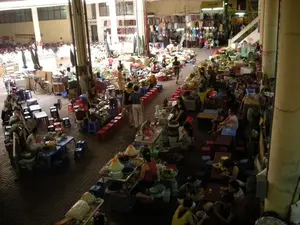 Đây là khung cảnh bên trong một khu chợ có mái che ở Hà Nội, không gian rất rộng lớn và đông đúc. Hoạt động chính là buôn bán và ăn uống. Ở phía trước và khu vực trung tâm là dãy các hàng quán ăn uống. Mọi người đang ngồi trên những chiếc ghế nhựa thấp màu đỏ và xanh dương, quây quần quanh những chiếc bàn thấp để ăn các món ăn được phục vụ trong bát. Trên các quầy hàng, có nhiều nồi, xoong và các nguyên liệu đã được chuẩn bị sẵn. Ở phía sau là khu vực bán hàng hóa khác, bao gồm các sạp trái cây và quần áo được treo trên giá. Có một cầu thang dẫn lên tầng trên của khu chợ. Nền nhà được lát gạch. Một tấm biển có chữ "VE SINH" (Nhà vệ sinh) có thể được nhìn thấy ở phía xa. Không khí trong chợ rất nhộn nhịp và sầm uất, phản ánh sinh hoạt thường ngày tại một khu chợ truyền thống của Việt Nam.。（日本語訳）。これはハノイのある屋根付き市場の内部の光景です。空間は非常に広く、混雑しています。主な活動は売買と飲食です。手前と中央エリアには飲食店の列が並んでいます。人々は赤と青の低いプラスチックの椅子に座り、低いテーブルを囲んで、お椀で提供される料理を食べています。屋台の上には、たくさんの鍋やフライパン、そして下準備された食材があります。後方には、果物の屋台やラックに掛けられた衣類を含む、他の商品を売るエリアがあります。市場の上階へ続く階段があります。床はタイル張りです。遠くに「VE SINH」（トイレ）と書かれた看板が見えます。市場の中の雰囲気は非常に賑やかで活気があり、ベトナムの伝統的な市場での日常生活を反映しています。