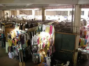 Trên một tấm bảng đen màu xanh lá cây có ghi số '6'. Bên dưới có một số chữ viết bằng phấn nhưng không thể đọc rõ.。Đây là khung cảnh bên trong một khu chợ bán quần áo trong nhà, có thể là một khu chợ đầu mối. Khu chợ có kết cấu nhiều tầng với các cột bê tông và dầm nhà lộ ra. Tầng dưới là các gian hàng quần áo san sát nhau, trong khi khu vực tầng trên có vẻ trống trải hơn với các khung kệ rỗng, có thể đang được sửa chữa hoặc dùng làm kho chứa hàng.。Các gian hàng trưng bày rất nhiều quần áo nữ, chủ yếu là áo kiểu và váy, được treo trên các giá kim loại và mặc cho ma-nơ-canh. Quần áo có nhiều màu sắc và kiểu dáng. Một người phụ nữ đang đứng giữa một gian hàng, có thể là người bán hàng đang sắp xếp đồ hoặc khách hàng đang lựa chọn. Có một chiếc xe máy màu trắng và một chiếc xe đạp dựng gần một gian hàng. Nhiều túi sọc lớn bằng nhựa, thường dùng để đựng hàng, được đặt trên sàn nhà. Khung cảnh cho thấy hoạt động buôn bán quần áo tại một khu chợ ở Hà Nội.。（日本語訳）。緑色の黒板に「6」という数字が書かれています。その下にはチョークで書かれた文字がありますが、はっきりと読むことはできません。これは屋内の衣料品市場の光景で、おそらく卸売市場でしょう。市場は多層構造で、コンクリートの柱と梁がむき出しになっています。下の階には衣料品店が密集していますが、上の階は空の棚が並び、よりがらんとしているように見え、おそらく改装中か倉庫として使われているのでしょう。各店舗には、主にブラウスやスカートといった多くの婦人服が展示されており、金属製のラックに掛けられたり、マネキンに着せられたりしています。服は色もデザインも様々です。一人の女性が店の真ん中に立っており、商品を整理している店員か、商品を選んでいる客かもしれません。ある店の近くに白いバイクと自転車が1台ずつ停められています。商品を運ぶのによく使われる、大きな縞模様のビニール袋が床にたくさん置かれています。この光景は、ハノイの市場での衣料品販売の様子を示しています。