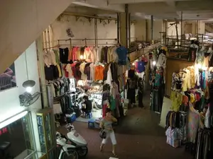 Bức ảnh chụp từ trên cao nhìn xuống một khu chợ quần áo trong nhà, có khả năng là một khu vực bên trong một khu chợ lớn ở Hà Nội. Không gian chợ có vẻ cũ với các cột bê tông, trần nhà và hệ thống đường ống lộ ra ngoài. Hoạt động chính là buôn bán và mua sắm quần áo. Rất nhiều quần áo, váy và áo sơ mi đủ màu sắc được treo dày đặc trên các giá và sào kim loại ở nhiều tầng khác nhau. Có một vài người trong ảnh: một người phụ nữ đang đi bộ ở lối đi chính, một vài người khác đang ngồi tại một quầy hàng trên những chiếc ghế đẩu nhựa màu xanh, có thể là người bán và khách mua hàng. Các vật thể đáng chú ý bao gồm vô số quần áo, ma-nơ-canh, một chiếc xe máy tay ga màu trắng đỗ ở phía trước bên trái, một chiếc quạt treo tường, và các loại đèn chiếu sáng. Trên tường có một tấm áp phích nhưng không thể đọc rõ nội dung.。（日本語訳）。上から見下ろした屋内の衣料品市場の写真で、ハノイの大きな市場内の一角である可能性が高い。市場の空間は、コンクリートの柱、天井、むき出しの配管があり、古びた印象を受ける。主な活動は衣料品の売買と買い物である。様々な色の服、ワンピース、シャツが、何段にもわたる金属製のラックやポールにびっしりと掛けられている。写真には数人の人物が写っている。メイン通路を歩いている一人の女性、青いプラスチック製のスツールに座って屋台にいる他の数人、おそらく売り手と買い手だろう。目立つ物としては、無数の衣類、マネキン、左手前に駐車された白いスクーター、壁掛け扇風機、そして様々な種類の照明器具が含まれる。壁にはポスターが一枚あるが、内容ははっきりと読み取れない。