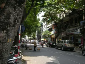 Bối cảnh là một con phố rợp bóng cây ở Hà Nội. Hai bên đường là những dãy nhà ống nhiều tầng san sát nhau, đặc trưng của kiến trúc đô thị Việt Nam, với tầng trệt dường như là các cửa hàng. Có nhiều biển hiệu quảng cáo trên các tòa nhà nhưng chữ viết bị mờ, khó đọc, tuy nhiên có thể nhận ra một biển hiệu có chữ 'bank'. Hệ thống dây điện chằng chịt vắt ngang phía trên.。Hoạt động chính trong ảnh là cảnh sinh hoạt đời thường trên phố. Nổi bật là hình ảnh hai người phụ nữ đội nón lá, đang gánh hàng rong bằng đòn gánh với hai chiếc thúng tre đi bộ dưới lòng đường. Đây là hình ảnh quen thuộc của những người bán hàng rong. Xung quanh, xe máy và ô tô đang đỗ dọc hai bên đường và di chuyển trên phố. Một vài người khác cũng xuất hiện trong khung cảnh, người thì ngồi trên vỉa hè, người thì đi xe máy.。Các vật thể đáng chú ý bao gồm những cây cổ thụ lớn với thân cây xù xì, một chiếc xe máy đang đỗ trên vỉa hè, và một cột biển báo giao thông sọc đỏ trắng.。（日本語訳）。背景はハノイの緑豊かな並木道です。道の両側には、ベトナムの都市建築に特徴的な、1階が店舗になっていると思われる多層階のチューブハウスが密集しています。建物には多くの広告看板がありますが、文字はぼやけていて読みにくいですが、「bank」という文字のある看板が一つ認識できます。頭上には電線が複雑に張り巡らされています。写真の中の主な活動は、路上での日常の生活風景です。際立っているのは、ノンラーをかぶった二人の女性が、天秤棒と二つの竹かごで露天商いをしながら車道を歩いている姿です。これは露天商のよく見られる光景です。周りでは、バイクや車が道の両側に沿って駐車していたり、通りを移動していたりします。他にも数人が風景の中に現れており、歩道に座っている人や、バイクに乗っている人がいます。注目すべき物体には、ごつごつした幹を持つ大きな古木、歩道に駐車している一台のバイク、そして赤と白の縞模様の交通標識のポールが含まれます。