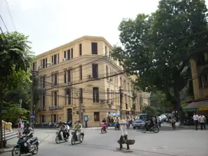 Bối cảnh là một góc phố tại Hà Nội với giao thông đông đúc. Một tòa nhà lớn, nhiều tầng, sơn màu vàng mang phong cách kiến trúc Pháp chiếm phần lớn khung hình. Trên đường, nhiều người đang điều khiển xe máy và đều đội mũ bảo hiểm. Nổi bật giữa dòng xe là hình ảnh một người phụ nữ đội nón lá đang gánh hàng rong đi bộ qua đường. Bên lề đường có nhiều cây xanh cổ thụ và một hệ thống dây điện chằng chịt vắt ngang phía trên. Có một số người đi bộ trên vỉa hè và một vài chiếc ô tô cũng xuất hiện trong ảnh. Có một biển báo đường phố màu xanh ghi chữ 'PHỐ TRÀNG THI'.。（日本語訳）。背景は交通量の多いハノイのある街角です。フランス建築様式の黄色に塗られた大きな多層階の建物が、構図の大部分を占めています。路上では、多くの人々がバイクを運転しており、全員がヘルメットを着用しています。車の流れの中でひときわ目立つのは、ノンラーをかぶった女性が天秤棒を担いで道を歩いて渡っている姿です。道端には多くの古い緑の木々があり、頭上には複雑に絡み合った電線が張り巡らされています。「PHỐ TRÀNG THI」と書かれた青い道路標識があります。