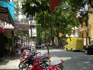 Đây là hình ảnh một con phố ở Hà Nội vào một ngày nắng. Bên trái là dãy cửa hàng san sát nhau, phía trước có mái hiên. Nhiều xe máy được dựng trên vỉa hè. Một vài người dân đang ngồi trên những chiếc ghế nhựa thấp để nghỉ ngơi hoặc trò chuyện. Có các biển hiệu của cửa hàng, trong đó có một biển hiệu lớn ghi "PS" và địa chỉ "50 HÀNG THIẾC". Một biển hiệu khác phía dưới quảng cáo món "LẨU". Dưới lòng đường, có một chiếc xe tải nhỏ màu vàng đang đậu, trên thân xe có chữ "Snackcar" và địa chỉ trang web "www.snackcar.vn". Phía sau chiếc xe tải là một chiếc ô tô màu đen. Con phố có nhiều cây xanh và các tòa nhà nhiều tầng với kiến trúc đa dạng, một số tòa nhà được sơn màu vàng đặc trưng. Hệ thống dây điện chằng chịt vắt ngang qua đường là một hình ảnh quen thuộc. Phía xa có thể thấy các phương tiện khác đang lưu thông.。（日本語訳）。これは晴れた日のハノイのある通りの写真です。左側には店が立ち並び、その前には軒があります。多くのバイクが歩道に停められています。何人かの人々が低いプラスチックの椅子に座って休憩したり、おしゃべりをしたりしています。店の看板があり、その中には「PS」と住所「50 HÀNG THIẾC」と書かれた大きな看板があります。その下にある別の看板は「LẨU」（鍋料理）を宣伝しています。道路には黄色い小さなトラックが停まっており、車体には「Snackcar」という文字とウェブサイトのアドレス「www.snackcar.vn」が書かれています。トラックの後ろには黒い乗用車がいます。通りには多くの緑の木々と、多様な建築様式の多層階の建物があり、いくつかの建物は特徴的な黄色に塗られています。複雑に絡み合った電線が通りを横切っているのは、おなじみの光景です。遠くには他の乗り物が通行しているのが見えます。