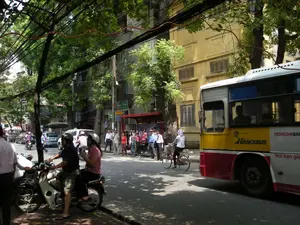 Bối cảnh là một con phố ở Hà Nội vào một ngày nắng. Con phố rợp bóng cây xanh và có nhiều phương tiện giao thông qua lại. Ở bên phải, một chiếc xe buýt lớn màu vàng, đỏ và trắng đang dừng đỗ. Trên thân xe buýt có các dòng chữ 'HANOIBUS', 'TECHCOMBANK' và một phần của dòng chữ 'Nơi bán vé...'. Phía trên kính chắn gió của xe buýt có thể thấy chữ 'DAEWOO'. Một nhóm người đang đứng đợi ở nhà chờ xe buýt màu đỏ. Một người phụ nữ mặc áo trắng, trùm đầu chống nắng, đang đi xe đạp trên đường. Ở phía trước, một cặp đôi (một nam, một nữ) đang ngồi trên một chiếc xe máy, cả hai đều đội mũ bảo hiểm. Nhiều người khác đang đi bộ trên vỉa hè và các phương tiện khác như ô tô, xe máy đang lưu thông trên đường. Phía sau là những tòa nhà nhiều tầng, trong đó có một tòa nhà màu vàng nổi bật với kiến trúc cửa sổ có chớp. Phía trên đường là một hệ thống dây điện và dây cáp chằng chịt, đặc trưng của cảnh quan đô thị Việt Nam.。（日本語訳）。舞台は晴れた日のハノイのある通りです。通りは緑の木々で覆われ、多くの交通車両が行き交っています。右側には、黄色、赤、白の大きなバスが停車しています。バスの車体には「HANOIBUS」、「TECHCOMBANK」という文字と、「乗車券販売所...」という文字の一部が見えます。バスのフロントガラスの上には「DAEWOO」という文字が見えます。赤いバス停で数人のグループが待っています。白いシャツを着て、日差しを避けるために頭を覆った女性が、道路を自転車で走っています。前方では、カップル（男性1人、女性1人）がバイクに乗っており、2人ともヘルメットをかぶっています。他にも多くの人々が歩道を歩いており、車やバイクなどの他の車両が道路を走行しています。後方には多層階の建物があり、その中には鎧戸のある窓の建築が特徴的な、目立つ黄色い建物があります。道路の上には、ベトナムの都市景観に特徴的な、複雑に絡み合った電線とケーブルのシステムがあります。