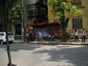 Đây là hình ảnh một con phố tại Hà Nội, có một nhà chờ xe buýt với mái che màu đỏ nằm bên vỉa hè. Nhiều người đang đứng và ngồi tại khu vực nhà chờ, dường như đang đợi xe buýt. Trong số họ có một người phụ nữ đội nón lá. Gần đó, một vài người trẻ mặc áo sơ mi trắng đang đứng trên vỉa hè. Phía sau là các tòa nhà nhiều tầng và cây xanh. Một chiếc ô tô màu trắng đang đỗ ở lề đường. Bên trong nhà chờ có một tấm biển quảng cáo lớn, có thể nhận ra logo của thương hiệu "OMO". Phía trên một cửa hàng có tấm biển hiệu màu xanh lá cây nhưng chữ viết không thể đọc rõ. Trên một cây cột gần đó cũng có một biển báo màu vàng nhưng nội dung không thể nhận dạng được.。（日本語訳）。これはハノイのある通りの画像です。歩道脇に赤い屋根のバス停があります。多くの人々がバス停のエリアで立ったり座ったりしており、バスを待っているようです。その中にはノンラーをかぶった女性が一人います。近くでは、白いシャツを着た数人の若者が歩道に立っています。後方には高層ビルと緑の木々があります。一台の白い車が路肩に駐車しています。バス停の中には大きな広告看板があり、「OMO」というブランドのロゴが認識できます。ある店の上には緑色の看板がありますが、文字ははっきりと読み取れません。近くの柱にも黄色い標識がありますが、その内容は識別できません。