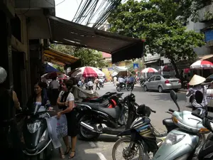 Đây là một cảnh đường phố nhộn nhịp ở Hà Nội. Con đường có nhiều người và xe cộ qua lại, chủ yếu là xe máy. Nhiều xe máy được đỗ dọc vỉa hè và dưới lòng đường, bên cạnh một chiếc ô tô màu bạc. Ở phía trước, hai người phụ nữ đang đứng cạnh một chiếc xe máy, một người cầm một chiếc túi mua sắm. Trong bối cảnh, có nhiều người khác đang đi bộ hoặc lái xe, và một người đang đội nón lá. Các cửa hàng ven đường có mái hiên và những chiếc ô lớn để che nắng. Có thể nhìn thấy chữ 'HABECO' và 'BIA HÀ NỘI' trên những chiếc ô này. Một biển hiệu cửa hàng ở phía xa có chữ '... LAN CHÍN'. Phía trên đường phố là một mạng lưới dây điện chằng chịt. Một chiếc xe máy ở gần có nhãn hiệu 'YAMAHA'.。（日本語訳）。これはハノイの賑やかな街の風景です。道路には多くの人々や乗り物が行き交っており、主にバイクです。多くのバイクが歩道沿いや車道に、銀色の自動車の隣に駐車されています。手前では、二人の女性がバイクの隣に立っており、一人は買い物袋を持っています。背景には、歩いていたり運転していたりする他の多くの人々がおり、一人はノンラーをかぶっています。道端の店には日よけの軒や大きな傘があります。これらの傘には「HABECO」と「BIA HÀ NỘI」の文字が見えます。遠くにある店の看板には「... LAN CHÍN」という文字があります。通りの上には、複雑に絡み合った電線の網があります。近くにあるバイクには「YAMAHA」のブランド名が付いています。
