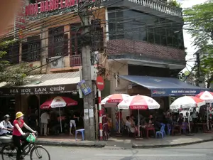 Chữ viết được ghi lại:。*   Trên tòa nhà có biển hiệu lớn màu đỏ: "NHÀ HÀNG LAN TUẤN".。*   Biển hiệu của quán ở tầng trệt: "BIA HƠI LAN CHÍN" và "NHẬN ĐẶT TIỆC, SINH NHẬT, HỘI NGHỊ".。*   Trên mái hiên màu xanh có logo "PEPSI" và dòng chữ "BIA HƠI LAN CHÍN".。*   Biển báo tên đường màu xanh gắn trên cột điện: "PHỐ HÀNG TRE".。*   Trên những chiếc ô che nắng có logo của "BIA HÀ NỘI" và "HABECO".。*   Một biển báo giao thông hình tròn màu đỏ (biển cấm).。Mô tả cảnh quan và hoạt động:。*   **Địa điểm:** Khung cảnh diễn ra tại một góc phố ở Hà Nội, có thể là trên phố Hàng Tre như trên biển báo. Đây là một quán bia hơi vỉa hè, một hình thức kinh doanh và sinh hoạt văn hóa đặc trưng. Các tòa nhà là dạng nhà ống nhiều tầng san sát nhau, phía trước có mạng lưới dây điện và cáp viễn thông chằng chịt.。*   **Hoạt động:** Nhiều người (chủ yếu là đàn ông) đang ngồi tụ tập tại các bàn nhựa thấp trên vỉa hè để uống bia hơi, ăn uống và trò chuyện. Đây là một hoạt động giao lưu xã hội phổ biến. Trên đường, một người phụ nữ đội mũ vàng, đeo khẩu trang đang đi xe đạp. Có cả người đang đi xe máy, cho thấy không khí tấp nập của đường phố.。*   **Vật thể:** Các vật thể nổi bật bao gồm những chiếc bàn và ghế nhựa thấp màu xanh, đỏ; những chiếc ô lớn của hãng Bia Hà Nội (Habeco) dùng để che nắng cho khách; một cột điện lớn với nhiều dây điện; xe đạp và xe máy đang lưu thông. Tòa nhà có kiến trúc kiểu cũ với cửa sổ chớp bằng gỗ.。（日本語訳）。記録された文字：。*   建物の上にある大きな赤い看板：「NHÀ HÀNG LAN TUẤN」（ラン・トゥアン・レストラン）。*   1階の店の看板：「BIA HƠI LAN CHÍN」（ラン・チン生ビール）と「NHẬN ĐẶT TIỆC, SINH NHẬT, HỘI NGHỊ」（パーティー、誕生日、会議の予約受付）。*   青い庇の上には「PEPSI」のロゴと「BIA HƠI LAN CHÍN」の文字。*   電柱に取り付けられた青い通りの名前の標識：「PHỐ HÀNG TRE」（ハンチェ通り）。*   日よけの傘の上には「BIA HÀ NỘI」（ハノイビール）と「HABECO」のロゴ。*   一つの赤い円形の交通標識（禁止標識）。景観と活動の説明：。*   **場所：** ハノイのある街角の光景で、標識にあるようにハンチェ通りである可能性が高い。これは歩道のビアホイ店であり、特徴的なビジネス形態および文化的な生活様式である。建物は多層のチューブハウス（間口が狭く奥行きが深い家）が密集しており、正面には電線や通信ケーブルが複雑に張り巡らされている。*   **活動：** 多くの人々（主に男性）が歩道上の低いプラスチック製のテーブルに集まって座り、ビアホイを飲み、食事をし、おしゃべりをしている。これは一般的な社会交流活動である。路上では、黄色い帽子をかぶり、マスクをした女性が自転車に乗っている。バイクに乗っている人もおり、通りの賑やかな雰囲気を示している。*   **物体：** 目立つ物体には、青や赤の低いプラスチック製のテーブルと椅子、客の日よけに使われるハノイビール社（Habeco）の大きな傘、多くの電線がついた大きな電柱、通行中の自転車やバイクが含まれる。建物は木製の鎧戸窓を持つ古い様式の建築である。