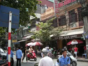 Bức ảnh ghi lại cảnh một con phố nhộn nhịp và đông đúc ở Hà Nội. Không khí tấp nập với nhiều người và xe cộ qua lại. Các tòa nhà là kiểu nhà ống, cao tầng, san sát nhau, với hệ thống dây điện và cáp viễn thông chằng chịt, một hình ảnh đặc trưng của các đô thị Việt Nam.。Hoạt động chính là sinh hoạt đường phố. Nhiều người đang di chuyển bằng xe máy, một số đang đi bộ. Có một quán bia hơi ở bên phải, nơi mọi người có thể đang ngồi ăn uống dưới những chiếc ô. Một người đàn ông mặc đồng phục màu xanh dương đang ngồi trên xe máy. Một người phụ nữ mặc áo đen đang đi bộ trên đường.。Các vật thể đáng chú ý bao gồm rất nhiều xe máy, một cột biển báo giao thông sơn sọc đỏ trắng ở phía trước bên trái, và các biển hiệu của cửa hàng.。Các dòng chữ có thể đọc được trên các biển hiệu bao gồm:。- Biển hiệu quán ăn: "BIA HƠI LAN CHÍN", "NHẬN ĐẶT TIỆC, SINH NHẬT, HỘI NGHỊ", và địa chỉ "15B TRẦN QUANG KHẢI".。- Biển hiệu lớn hơn ở tầng trên: "NHÀ HÀNG LAN...".。- Biển tên đường màu xanh: "PHỐ HÀNG TRE".。- Trên một chiếc ô: "BIA HÀ NỘI" và "HABECO".。- Trên mặt tiền một tòa nhà kiến trúc cổ màu vàng có khắc số "1923".。（日本語訳）。写真は、ハノイの賑やかで混雑した通りの光景を捉えています。多くの人々や車両が行き交い、活気のある雰囲気です。建物は縦に細長い高層の様式で、互いに密集しており、電線や通信ケーブルが複雑に絡み合っている様子は、ベトナムの都市の特徴的な光景です。主な活動は路上での生活風景です。多くの人々がバイクで移動しており、何人かは歩いています。右側にはビアホイの店があり、人々がパラソルの下で飲食しているのかもしれません。青い制服を着た男性がバイクに座っています。黒い服を着た女性が路上を歩いています。注目すべき物体には、非常に多くのバイク、左手前にある赤と白の縞模様に塗られた交通標識の柱、そして店の看板が含まれます。看板で読み取れる文字には、以下のものが含まれます：。- 飲食店の看板：「BIA HƠI LAN CHÍN」、「NHẬN ĐẶT TIỆC, SINH NHẬT, HỘI NGHỊ」、そして住所「15B TRẦN QUANG KHẢI」。- 上の階のより大きな看板：「NHÀ HÀNG LAN...」。- 青い通りの名前の標識：「PHỐ HÀNG TRE」。- 一つのパラソルに：「BIA HÀ NỘI」と「HABECO」。- 黄色い古い建築様式の建物のファサードに、数字「1923」が刻まれています。