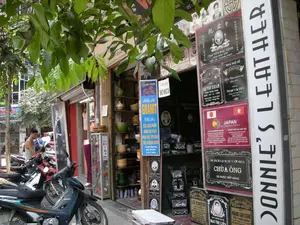 ハノイの街並みを撮影した写真で、ある店のファサード（正面）に焦点を当てています。これは石彫の専門店で、特に墓石や各種記念碑、石の看板などを製作しています。**風景と活動の詳細：**。*   **場所：** ある通りの歩道にある店。店の前のスペースは、完成した石製品の展示とバイクの駐車に使われています。*   **活動：** 主な活動は、石の美術工芸品の販売と展示です。多くのバイクが駐車していることから、ここは人通りの多い地域であることがわかります。一人の男性がバイクの近くに立っています。*   **物体：**。*   **石碑：** 多くの花崗岩製の墓石や記念碑が、見本として店の外に立てられています。これらの石碑には、肖像画、名前、年月日、その他の情報が刻まれています。*   **看板：** 多くの看板や文字があります。白い縦長の看板には「DONNIE\'S LEATHER」と書かれています。青い看板は店のサービスを宣伝しています：「花崗岩の彫刻を専門に請け負います」、「お客様のご要望に応じたデザインで石製品を製作します。高品質。美術的、高級。」。*   **バイク：** 主にカブタイプのバイクが一列に、歩道にぎっしりと停められています。*   **その他の商品：** 隣の店には、陶磁器や漆器（椀、壺）のような製品を展示する棚があります。*   **樹木：** 前方にある木の緑の葉が、通りに木陰を作っています。**記録された文字の内容：**。*   **見本の墓石に書かれた内容：**。*   「NGUYỄN ĐẮC TỈNH 生年1920 没：18-9 庚申（かのえさる）」。*   「ÔNG SẢI VĂN KHOẢN 生年1950 没：1982年6月29日 故郷：ĐỒNG YÊN - LẠC THỦY...」。*   「LÊ CHIÊU」、「MAI THỊ LIÊN」、「HOÀNG VĂN CHÂN 字（あざな）：TRẦN ĐOÀN」。*   **その他の看板や見本石碑に書かれた内容：**。*   「ファム一族の祖先を祀る教会堂」。*   「オン寺の歴史文化遺跡はランク付けされました」。*   日本とベトナムの国旗が描かれた赤い見本看板には、次のように書かれています：「JAPAN」、「日本政府及び日本国民の援助によるベトナム・日本友好小学校...」