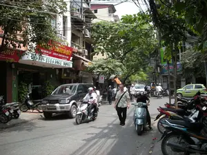 Bức ảnh ghi lại cảnh một con phố nhộn nhịp ở Hà Nội vào ban ngày. Các dòng chữ trên biển hiệu có thể đọc được bao gồm: "HANOI ELEGANCE HOTEL", "KHÁCH SẠN DU LỊCH", "VÀNG BẠC ĐÁ QUÝ", "JEWELRY - GEMSTONE PICTURE", và một biển hiệu có logo tách cà phê ghi chữ "Cafe". Một biển số nhà ghi số "8".。Đây là một con phố với những tòa nhà nhiều tầng san sát nhau, đặc trưng của kiến trúc đô thị Việt Nam. Tầng trệt của các tòa nhà là mặt bằng kinh doanh, bao gồm một khách sạn và các cửa hàng khác. Cây xanh được trồng dọc hai bên đường, tạo ra không gian râm mát. Một đặc điểm nổi bật là hệ thống dây điện và dây cáp chằng chịt vắt ngang phía trên đường.。Hoạt động chính diễn ra trên phố là giao thông đi lại. Nhiều người đang điều khiển xe máy (xe ga và xe số), phần lớn đều đội mũ bảo hiểm. Một chiếc ô tô SUV màu sẫm đang đỗ bên lề đường, và nhiều xe máy khác cũng được dựng trên vỉa hè. Ở phía xa, có thể thấy một chiếc taxi màu xanh lá cây và các phương tiện khác đang lưu thông. Một vài người đang đi bộ trên đường, trong đó có một người đàn ông nước ngoài. Khung cảnh phản ánh nhịp sống đô thị sôi động và thường nhật.。（日本語訳）。写真はハノイの昼間の賑やかな通りの光景を記録したものです。看板に書かれている読み取れる文字には、「HANOI ELEGANCE HOTEL」、「KHÁCH SẠN DU LỊCH（観光ホテル）」、「VÀNG BẠC ĐÁ QUÝ（金銀宝石）」、「JEWELRY - GEMSTONE PICTURE（ジュエリー - 宝石画）」、そして「Cafe」という文字とコーヒーカップのロゴがある看板が含まれます。ある番地表示には「8」という数字が書かれています。これは、ベトナムの都市建築に特徴的な、多層階の建物が密集している通りです。建物の1階は商業スペースで、ホテルやその他の店舗が含まれています。通りの両側には街路樹が植えられており、日陰の空間を作り出しています。際立った特徴は、通りを横切るように張り巡らされた、複雑な電線とケーブルのシステムです。通りで行われている主な活動は交通です。多くの人々がバイク（スクーターとマニュアル車）を運転しており、そのほとんどがヘルメットを着用しています。濃い色のSUV車が路肩に駐車しており、他の多くのバイクも歩道に停められています。遠くには、緑色のタクシーやその他の車両が通行しているのが見えます。数人が道を歩いており、その中には外国人男性が一人います。この光景は、活気に満ちた日常的な都市の生活リズムを反映しています。