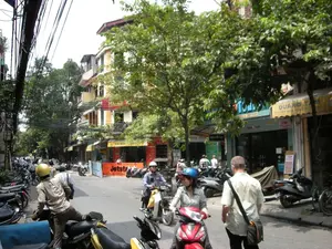 Bối cảnh là một con phố nhộn nhịp ở Hà Nội, có thể là một khu vực có nhiều khách du lịch. Hai bên đường là những tòa nhà ống cao tầng đặc trưng với các cửa hàng ở tầng trệt, và có nhiều cây xanh. Hoạt động chính là giao thông đường phố, với rất nhiều người đang di chuyển bằng xe máy và một số người đi bộ. Nhiều người đi xe máy đội mũ bảo hiểm. Có thể thấy một người đàn ông đội mũ bảo hiểm màu vàng đang chuẩn bị lên xe, một phụ nữ đội mũ bảo hiểm màu xanh đang ngồi trên xe máy màu đỏ, và một người đàn ông khác đang đi bộ trên đường. Một đặc điểm nổi bật là hệ thống dây điện và dây cáp chằng chịt ở phía trên. Các biển hiệu có thể đọc được bao gồm một tấm biển quảng cáo màu cam có chữ 'Jetstar', một mái hiên màu vàng ghi 'HA NOI TOUR...', một biển hiệu màu xanh lam có chữ 'Tou...' và 'TRAVELLED', và một mái hiên khác ghi 'QUANG T...'.。（日本語訳）。背景はハノイの賑やかな通りで、おそらく多くの観光客がいるエリアでしょう。通りの両側には、1階に店舗が入った特徴的な高層のチューブハウスが立ち並び、緑の木々も多く見られます。主な活動は道路交通で、多くの人々がバイクで移動し、何人かの歩行者もいます。バイクに乗っている多くの人がヘルメットを着用しています。黄色いヘルメットをかぶった男性がバイクに乗ろうとしているところ、青いヘルメットをかぶった女性が赤いバイクに座っているところ、そして別の男性が道を歩いているのが見えます。際立った特徴は、頭上にある絡み合った電線とケーブルのシステムです。読み取れる看板には、「Jetstar」という文字が入ったオレンジ色の広告看板、「HA NOI TOUR...」と書かれた黄色い日よけ、「Tou...」と「TRAVELLED」という文字が入った青い看板、そして「QUANG T...」と書かれた別の日よけなどがあります。