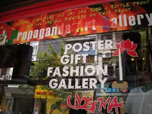 Bức ảnh chụp mặt tiền của một phòng trưng bày nghệ thuật trên một con phố ở Hà Nội. Trên tấm biển hiệu lớn màu đỏ ở phía trên có dòng chữ tiếng Anh: "CULTURE, LITERATURE AND ART ARE ALSO A BATTLE FRONT AND YOU ARE ALL FIGHTERS ON THAT FRONT", và tên phòng trưng bày là "Propaganda Art Gallery". Dán trên cửa kính là các chữ lớn màu trắng quảng cáo các mặt hàng của cửa hàng: "POSTER" (Áp phích), "GIFT" (Quà tặng), "FASHION" (Thời trang), "GALLERY" (Phòng trưng bày). Ở góc dưới có chữ "DOGMA" được thiết kế cách điệu màu đỏ. Trên kính còn có hình dán một bông hoa sen màu đỏ. Khung cảnh đường phố đối diện được phản chiếu qua cửa kính, cho thấy các tòa nhà, cây xanh và một mạng lưới dây điện chằng chịt. Có thể thấy một phần biển hiệu của các cửa hàng khác bị phản chiếu ngược, chẳng hạn như một tấm biển màu vàng có chữ "NHÀ HÀNG".。（日本語訳）。ハノイのある通りにあるアートギャラリーの正面を撮影した写真です。上部にある大きな赤い看板には、英語で「CULTURE, LITERATURE AND ART ARE ALSO A BATTLE FRONT AND YOU ARE ALL FIGHTERS ON THAT FRONT」（文化、文学、芸術もまた一つの戦線であり、あなた方はその戦線の戦士である）という文言と、「Propaganda Art Gallery」というギャラリー名が書かれています。ガラスのドアには、店の取扱商品を宣伝する大きな白い文字が貼られています：「POSTER」（ポスター）、「GIFT」（ギフト）、「FASHION」（ファッション）、「GALLERY」（ギャラリー）。下隅には、赤色で様式化された「DOGMA」という文字があります。ガラスには赤い蓮の花のステッカーも貼られています。向かいの通りの風景がガラスのドアに映り込んでおり、建物、緑の木々、そして複雑に絡み合った電線網が見えます。「NHÀ HÀNG」（レストラン）という文字が入った黄色い看板など、他の店の看板の一部が反転して映り込んでいるのが見えます。