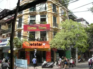 Bối cảnh là một con phố đô thị sầm uất ở Hà Nội, với các tòa nhà mang phong cách kiến trúc thuộc địa Pháp. Một mạng lưới dây điện và cáp viễn thông dày đặc, chằng chịt giăng mắc phía trên đường. Dưới lòng đường và trên vỉa hè, dòng người và xe cộ qua lại tấp nập.。**Thông tin chữ viết:**。- Biển hiệu màu đỏ lớn: "Hanoi Tours", bên dưới có dòng chữ "DỊCH VỤ DU LỊCH HÀ NỘI".。- Biển hiệu màu cam của đại lý vé máy bay: "Jetstar", "Đại lý 24 Hàng Bạc Tel: 049260809", "Jetstar.com", và "HA... TOURS".。- Biển hiệu ngân hàng bên trái: "GP-BANK".。- Biển quảng cáo di động màu xanh dương: "Pacific Airlines".。**Cảnh vật và hoạt động:**。- **Loại địa điểm:** Một con phố thương mại ở Hà Nội, cụ thể là phố Hàng Bạc, với các cửa hàng kinh doanh ở tầng trệt của các tòa nhà nhiều tầng.。- **Hoạt động cụ thể:** Người dân và khách du lịch (một số là người nước ngoài) đang đi bộ trên vỉa hè. Giao thông trên đường phố chủ yếu là xe máy và có cả xe đạp. Trước cửa hàng Jetstar, một người đàn ông đang đứng trên thang để làm việc, có thể là sửa chữa hoặc trang trí, trong khi một người khác giữ thang. Các hoạt động kinh doanh của ngân hàng và đại lý du lịch đang diễn ra.。- **Các vật thể trong ảnh:** Các tòa nhà cao tầng, nhiều xe máy (xe tay ga) đang đỗ và lưu thông, một cây xanh lớn, một cột điện, một chiếc thang, và nhiều biển hiệu quảng cáo. Một số người đi bộ đội mũ, trong đó có một người phụ nữ đội nón lá.。（日本語訳）。背景はハノイの賑やかな都市の通りで、フランス植民地時代の建築様式の建物が並んでいます。道路の上には、電線や通信ケーブルが密集し、複雑に張り巡らされています。車道と歩道では、人や車がひっきりなしに行き交っています。**書かれている情報：**。- 大きな赤い看板：「Hanoi Tours」、その下には「DỊCH VỤ DU LỊCH HÀ NỘI」という文字。- 航空券代理店のオレンジ色の看板：「Jetstar」、「Đại lý 24 Hàng Bạc Tel: 049260809」（代理店 24ハンバック通り 電話: 049260809）、「Jetstar.com」、そして「HA... TOURS」。- 左側の銀行の看板：「GP-BANK」。- 青い移動式広告看板：「Pacific Airlines」。**風景と活動：**。- **場所の種類：** ハノイの商業通り、具体的にはハンバック通りで、多層階の建物の1階に店舗が並んでいます。- **具体的な活動：** 地元の人々と観光客（一部は外国人）が歩道を歩いています。道路の交通は主にバイクで、自転車も見られます。Jetstarの店の前では、一人の男性がはしごに立って作業をしており（修理か装飾かもしれません）、その間、別の人がはしごを支えています。銀行や旅行代理店の営業活動が行われています。- **写真の中の物体：** 高層ビル、駐車したり走行したりしている多くのバイク（スクーター）、大きな緑の木一本、電柱一本、はしご一つ、そして多くの広告看板。歩行者の中には帽子をかぶっている人もおり、その中にはノンラーをかぶった女性が一人います。
