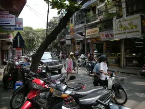 Bức ảnh ghi lại cảnh một con phố sầm uất và nhộn nhịp ở Hà Nội. Dựa trên các biển hiệu, đây có thể là phố Hàng Bạc, một khu vực kinh doanh sầm uất. Các biển hiệu có thể đọc được bao gồm: 'Công ty TNHH Vàng Bạc Đá Quý Diệp Kim', 'DOANH NGHIỆP VÀNG BẠC ĐỨC KHA', 'KIM LONG', và dịch vụ 'ĐỔI NGOẠI TỆ' tại địa chỉ '130 HÀNG BẠC'. Ngoài ra còn có biển hiệu 'HOTEL TẠI HÀ NỘI' trên phố Bà Trưng và 'CD & DVD'. Hoạt động chính trên phố là giao thông, với rất nhiều xe máy đang di chuyển và đậu san sát bên lề đường. Mọi người đang đi lại, một số lái xe, một số đi bộ. Có thể thấy một người phụ nữ mặc đồ màu hồng đang đi bộ và một người khác đội nón lá ở phía xa. Các vật thể nổi bật bao gồm nhiều loại xe máy, một chiếc ô tô màu đen, các tòa nhà kiểu nhà ống đặc trưng và một cây lớn mọc trên vỉa hè với hệ thống dây điện phức tạp phía trên.。（日本語訳）。写真はハノイの賑やかで活気のある通りの光景を捉えています。看板から判断すると、ここは賑やかな商業地区であるハンバック通りの可能性があります。読み取れる看板には、「Công ty TNHH Vàng Bạc Đá Quý Diệp Kim」、「DOANH NGHIỆP VÀNG BẠC ĐỨC KHA」、「KIM LONG」、そして住所「130 HÀNG BẠC」にある「ĐỔI NGOẠI TỆ」（外貨両替）サービスが含まれます。その他にも、バーチュン通りには「HOTEL TẠI HÀ NỘI」（ハノイのホテル）と「CD & DVD」の看板もあります。通りでの主な活動は交通で、非常に多くのバイクが移動したり、道路脇にびっしりと駐車したりしています。人々は行き交っており、一部は運転し、一部は歩いています。ピンク色の服を着た女性が歩いているのが見え、遠くにはノンラーをかぶった別の人がいます。目立つ物には、多くの種類のバイク、一台の黒い自動車、特徴的なチューブハウス様式の建物、そして上部に複雑な電線システムがある歩道に生えた大きな木が含まれます。