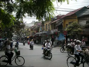 Bức ảnh chụp một con phố đông đúc ở Hà Nội, có khả năng là trong khu Phố Cổ. Khung cảnh đặc trưng với những ngôi nhà ống nhiều tầng, san sát nhau, kiến trúc có phần cũ kỹ. Tầng trệt của các ngôi nhà được sử dụng làm cửa hàng, bày bán các mặt hàng như quần áo. Trên đường, dòng người và phương tiện qua lại tấp nập, chủ yếu là xe máy và xe đạp. Nhiều người điều khiển xe máy đội mũ bảo hiểm. Nổi bật trong ảnh là một người phụ nữ đội nón lá truyền thống đang đi xe đạp. Phía trên là hệ thống dây điện, dây cáp chằng chịt. Cây xanh tỏa bóng mát xuống một phần con đường. Về chữ viết, có thể đọc được biển hiệu "GUEST HOUSE" và "BIANCO". Ngoài ra còn có một biển báo giao thông hình tròn màu đỏ có vạch ngang màu trắng, là biển báo cấm đi vào. Các biển hiệu khác của cửa hàng có chữ nhưng không thể đọc rõ nội dung.。（日本語訳）。写真はハノイの混雑した通りを撮影したもので、おそらく旧市街の中だと思われます。多層階建てのチューブハウスが密集し、やや古風な建築様式が特徴的な風景です。家々の1階は店舗として利用され、衣類などの商品が陳列されています。路上では、人や乗り物がひっきりなしに行き交い、主にバイクや自転車です。バイクを運転する多くの人がヘルメットを着用しています。写真の中でひときわ目立つのは、伝統的なノンラーをかぶって自転車に乗っている女性です。上には、電線やケーブルが複雑に張り巡らされています。緑の木々が道路の一部に涼しい影を落としています。文字については、「GUEST HOUSE」と「BIANCO」という看板が読み取れます。その他、白い横線が入った赤色の円形の交通標識があり、これは進入禁止の標識です。他の店舗の看板にも文字がありますが、内容ははっきりと読み取れません。
