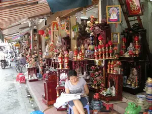 Khung cảnh chụp tại một con phố ở Hà Nội, nơi các cửa hàng ven đường bày bán rất nhiều đồ thờ cúng và vật phẩm tâm linh. Hàng hóa được trưng bày trên các kệ gỗ và tràn ra cả vỉa hè dưới mái hiên. Các sản phẩm nổi bật bao gồm nhiều loại bàn thờ gia tiên bằng gỗ với các kích cỡ khác nhau, đèn thờ hình hoa sen màu đỏ, chân nến, lư hương, các bức tượng Phật và Bồ Tát (như tượng Quan Âm), và các bức tranh thờ được lồng trong khung kính. Màu sắc chủ đạo của các vật phẩm là đỏ và vàng. Ở tiền cảnh, một người phụ nữ đang ngồi trên một chiếc ghế nhựa và đọc báo, có lẽ là người trông coi cửa hàng. Ở phía sau, trên lòng đường, có một người đội nón lá ngồi trên xe đạp và nhiều người khác đang đi lại bằng xe máy, tạo nên một không khí đường phố đặc trưng. Có một số biểu ngữ và tranh treo tường có chữ viết nhưng không thể đọc rõ nội dung.。（日本語訳）。ハノイのある通りで撮影された風景。そこでは道端の店が多くの祭祀用品やスピリチュアルな品物を販売している。商品は木製の棚に陳列され、軒下の歩道にまで溢れ出ている。目立つ商品には、様々なサイズの木製祖先祭壇、赤い蓮の花の形をした祭壇用の灯り、燭台、香炉、仏陀や菩薩の像（観音像など）、そしてガラスの額縁に入れられた祭壇画が含まれる。品物の主な色合いは赤と黄色である。前景には、一人の女性がプラスチックの椅子に座って新聞を読んでおり、おそらく店の番人だろう。後方、路上では、ノンラーをかぶった人が自転車に乗り、他の多くの人々がバイクで行き来しており、特徴的な通りの雰囲気を醸し出している。いくつかの横断幕や壁掛けの絵には文字が書かれているが、内容ははっきりと読み取れない。