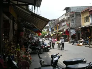 Bối cảnh là một con phố ở Hà Nội với những ngôi nhà ống đặc trưng. Hai bên đường là các cửa hàng, vỉa hè có rất nhiều xe máy đang đậu. Cửa hàng bên trái bày bán các vật phẩm trang trí màu vàng và đỏ, có thể là đồ vàng mã dùng trong thờ cúng. Trên đường, một người phụ nữ đội nón lá đang dắt một chiếc xe đạp. Những người khác đang đi bộ hoặc đi xe máy. Một lá cờ Việt Nam được treo dưới mái hiên của một cửa hàng. Có nhiều biển hiệu trên các tòa nhà nhưng không thể đọc rõ nội dung.。（日本語訳）。背景は、特徴的なチューブハウスが立ち並ぶハノイのある通りです。道の両側には店が並び、歩道にはたくさんのバイクが停まっています。左側の店は黄色と赤色の装飾品を陳列販売しており、それらは祭祀で使われる紙製の供物（冥器）かもしれません。路上では、ノンラーをかぶった一人の女性が自転車を押しています。他の人々は歩いていたり、バイクに乗っていたりします。ある店の軒下には、ベトナム国旗が一枚掲げられています。建物には多くの看板がありますが、その内容ははっきりと読み取れません。