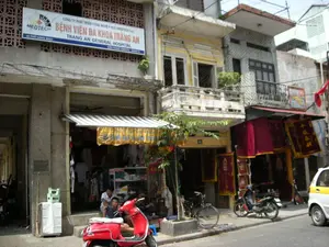 Bối cảnh là một con phố ở Hà Nội với những ngôi nhà ống san sát nhau. Trên tầng hai của một tòa nhà có một biển hiệu lớn với nội dung: "MEDTECH", "CÔNG TY PHÁT TRIỂN CÔNG NGHỆ Y HỌC (MEDTECH Co.)", "BỆNH VIỆN ĐA KHOA TRÀNG AN", "TRANG AN GENERAL HOSPITAL", "44 PHỐ HÀNG QUẠT", "ĐIỆN THOẠI: 8249274 - 8289273". Bên dưới là các cửa hàng đang hoạt động. Một cửa hàng bán quần áo có mái hiên sọc và một băng rôn màu vàng có chữ nhưng khó đọc rõ. Phía trước cửa hàng này, hai người đàn ông đang ngồi trên bậc thềm. Một cửa hàng khác bên cạnh treo bán nhiều tấm liễn, cờ phướn màu đỏ có thêu chữ Hán-Nôm màu vàng, có thể là vật phẩm dùng trong thờ cúng hoặc nghi lễ. Trên vỉa hè và lề đường có nhiều phương tiện giao thông đang đậu, bao gồm một chiếc xe máy tay ga màu đỏ ở phía trước, một chiếc xe máy khác, một chiếc xe đạp và một phần của một chiếc ô tô màu trắng vàng. Cảnh tượng thể hiện sinh hoạt và kinh doanh thường ngày trên một con phố.。（日本語訳）。背景は、細長い家々が密集するハノイのある通りです。ある建物の2階には、「MEDTECH」、「医療技術開発会社（MEDTECH Co.）」、「チャンアン総合病院」、「TRANG AN GENERAL HOSPITAL」、「ハンクアット通り44番地」、「電話：8249274 - 8289273」という内容の大きな看板があります。その下には営業中の店舗があります。ある衣料品店には縞模様の庇があり、文字が書かれているものの判読が難しい黄色い横断幕が掲げられています。この店の前では、2人の男性が階段に座っています。隣の別の店では、黄色いチュハン・ノム（漢字・チュノム）の文字が刺繍された多数の赤い対聯や幟が販売用に吊るされており、これらは祭祀や儀式で使われる品物かもしれません。歩道と路肩には、手前にある赤いスクーター1台、別のバイク1台、自転車1台、そして黄白色の自動車の一部を含む、多くの乗り物が駐車しています。この光景は、ある通りの日常的な生活と商売の様子を示しています。