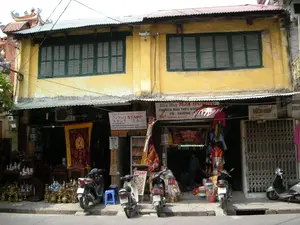 Bức ảnh chụp một con phố ở Hà Nội, có thể là trong khu Phố Cổ. Nổi bật là một tòa nhà hai tầng cũ sơn màu vàng. Tầng trên có các cửa sổ chớp bằng gỗ màu xanh lá cây. Tầng trệt là một dãy các cửa hàng kinh doanh với mái hiên bằng tôn.。Các biển hiệu có thể đọc được bao gồm:。1. Một cửa hàng khắc dấu với biển hiệu bằng nhiều ngôn ngữ: "THE REAL STAMPSHOP", "フックロイ STAMP" (Phúc Lợi STAMP), và "スタンプ はんこ" (con dấu).。2. Một nhà may có biển hiệu "NHÀ MAY MAY THÊU KHĂN", "CHUYÊN MAY THÊU KHĂN, CỜ - TRƯỜNG" tại địa chỉ "60A HÀNG QUẠT".。3. Một cửa hàng tên "BÍCH HỢP" ở số 60.。Hoạt động chính là kinh doanh tại các cửa hàng. Cửa hàng bên trái bán đồ cổ và đồ lưu niệm, trưng bày nhiều đồ đồng, đồ gốm sứ và một bức trướng thêu lớn. Cửa hàng khắc dấu và nhà may đang mở cửa, có người ở bên trong. Cửa hàng Bích Hợp thì đang đóng cửa bằng cửa cuốn kim loại. Một vài chiếc xe máy đang đậu trên vỉa hè. Phía trên là hệ thống dây điện chằng chịt, và ở góc xa bên trái có thể thấy một phần mái của một công trình kiến trúc truyền thống như đình hoặc chùa.。（日本語訳）。ハノイのある通りを撮影した写真で、おそらく旧市街の中でしょう。目立っているのは、黄色に塗られた古い2階建ての建物です。上の階には緑色の木製よろい戸の窓があります。1階はトタン屋根の軒先を持つ店舗が並んでいます。読み取れる看板には以下のものが含まれます：。1. 多言語の看板を掲げた印鑑彫刻店：「THE REAL STAMPSHOP」、「フックロイ STAMP」（Phúc Lợi STAMP）、そして「スタンプ はんこ」（印鑑）。2. 「NHÀ MAY MAY THÊU KHĂN」、「CHUYÊN MAY THÊU KHĂN, CỜ - TRƯỜNG」という看板を掲げた仕立て屋。住所は「60A HÀNG QUẠT」。3. 60番地にある「BÍCH HỢP」という名前の店。主な活動は店舗での商売です。左側の店は骨董品やお土産を販売しており、多くの銅製品、陶磁器、そして大きな刺繍の掛け軸を展示しています。印鑑彫刻店と仕立て屋は営業中で、中に人がいます。Bích Hợp店は金属製のシャッターで閉まっています。歩道には数台のバイクが停まっています。上方には複雑に絡み合った電線があり、左奥の角には亭や寺のような伝統的な建築物の一部の屋根が見えます。