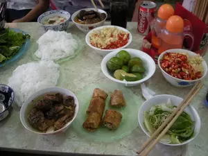 Hình ảnh ghi lại một bữa ăn tại một quán ăn, có khả năng là ở Hà Nội, với món đặc sản bún chả. Trên bàn ăn được bày biện thịnh soạn gồm nhiều món. Nổi bật là hai suất bún rối trắng tinh đặt trên đĩa. Có hai bát chả thịt lợn nướng ngâm trong nước mắm chua ngọt. Bên cạnh đó là một đĩa nem rán đã được cắt thành từng miếng nhỏ. Một đĩa rau sống gồm xà lách và các loại rau thơm khác dùng để ăn kèm. Các loại gia vị được chuẩn bị sẵn trong các bát nhỏ, bao gồm ớt tươi và tỏi băm, và một bát chanh tươi đã được cắt sẵn để vắt lấy nước. Về đồ uống, có một lon nước ngọt hiệu 'Coca-Cola' và một ly nước ngọt màu đen. Ngoài ra, trên bàn còn có các lọ đựng gia vị nắp cam và một hộp đựng đũa màu đỏ. Một vài người đang ngồi quanh bàn và thưởng thức bữa ăn.。（日本語訳）。写真は、おそらくハノイの食堂での、名物料理ブンチャーの食事を記録したものです。食卓には多くの料理が豪華に並べられています。目立っているのは、皿に盛られた真っ白なブン（米麺）の2人前です。甘酸っぱいヌクマム（魚醤）に浸された焼き豚チャーの入ったお椀が2つあります。その隣には、小さく切り分けられた揚げ春巻き（ネムザン）の皿があります。付け合わせ用の、レタスやその他の香草を含む生野菜の皿が1枚あります。生の唐辛子と刻みニンニクを含む各種薬味が小鉢に用意されており、また、絞るためのカットされた新鮮なライムの入った小鉢もあります。飲み物については、「コカ・コーラ」ブランドの炭酸飲料の缶が1つと、黒い炭酸飲料の入ったグラスが1つあります。その他、テーブルの上にはオレンジ色の蓋の調味料入れと、赤い箸立てがあります。数人がテーブルを囲んで座り、食事を楽しんでいます。
