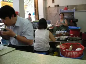 Bức ảnh chụp khung cảnh bên trong một quán ăn bình dân ở Hà Nội. Ở phía trước, một người đàn ông mặc áo sơ mi kẻ sọc đang ngồi ăn, tay cầm một chiếc bát nhỏ. Ngồi cạnh ông là một người phụ nữ mặc áo trắng, quay lưng về phía máy ảnh. Ở phía sau, trong khu vực có vẻ là bếp hoặc quầy chuẩn bị, một người phụ nữ khác đang ngồi. Không gian quán có các vật dụng như bàn ăn mặt đá, nhiều chai lọ gia vị, nồi lớn, một chiếc rổ nhựa màu đỏ, và một chiếc quạt điện. Nhìn qua cửa ra vào, có thể thấy một mái hiên sọc xanh lá cây và trắng. Bên dưới mái hiên có một tấm biển hiệu màu vàng chữ đỏ nhưng bị mờ và che khuất, chỉ có thể đọc được một phần chữ có thể là "CH... GÀ S...".。（日本語訳）。ハノイのある庶民的な食堂の内部の光景を撮影した写真です。手前には、ストライプのシャツを着た男性が座って食事をしており、手には小さな碗を持っています。彼の隣には、白い服を着た女性がカメラに背を向けて座っています。奥では、厨房か調理カウンターと思われるエリアに、別の女性が座っています。店内には、石の天板の食卓、たくさんの調味料の瓶、大きな鍋、赤いプラスチック製の籠、そして扇風機などの物があります。出入り口から外を見ると、緑と白のストライプの日よけが見えます。日よけの下には、黄色地に赤文字の看板がありますが、ぼやけて隠れており、「CH... GÀ S...」と思われる文字の一部しか読み取れません。