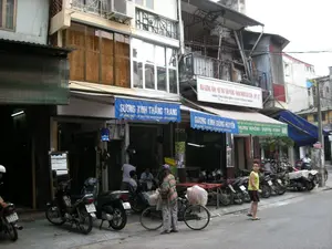 Bản ghi văn bản từ các biển hiệu:。1. Biển hiệu màu xanh dương bên trái: GƯƠNG KÍNH THẮNG TRANG, 69 HÀNG THIẾC - ĐT: 8247703-0912928689 - 0986969227.。2. Biển hiệu màu xanh dương ở giữa: GƯƠNG KÍNH DŨNG HUYỀN.。3. Biển hiệu lớn màu trắng ở trên: MAI GƯƠNG KÍNH - NỘI THẤT VĂN PHÒNG - KHUNG NHÔM CỬA CUỐN - CỬA THỦY LỰC. VẬN CHUYỂN ĐẾN CHÂN CÔNG TRÌNH.。4. Biển hiệu màu xanh lá và trắng bên phải: HÙNG NHÔM - GƯƠNG KÍNH.。Mô tả cảnh quan và hoạt động:。Bức ảnh chụp một con phố ở Hà Nội, có thể là phố Hàng Thiếc dựa trên địa chỉ trên biển hiệu. Đây là một khu phố thương mại với những ngôi nhà ống đặc trưng, mặt tiền được sử dụng làm cửa hàng. Các cửa hàng này chuyên về gương, kính, và khung nhôm. Nhiều xe máy được đậu dọc vỉa hè. Hoạt động trong ảnh là cảnh sinh hoạt đời thường: một người phụ nữ đang đẩy xe đạp chở hàng, một cậu bé đứng trên đường, và một vài người đàn ông đang ngồi trước cửa hàng. Bên trong một cửa hàng có một chiếc gương lớn được dựng lên.。（日本語訳）。看板の文字起こし：。1. 左側の青い看板：GƯƠNG KÍNH THẮNG TRANG、69 HÀNG THIẾC - 電話：8247703-0912928689 - 0986969227。2. 中央の青い看板：GƯƠNG KÍNH DŨNG HUYỀN。3. 上部の大きな白い看板：MAI GƯƠNG KÍNH - オフィス内装 - アルミフレーム・シャッタードア - 油圧ドア。工事現場まで輸送。4. 右側の緑と白の看板：HÙNG NHÔM - GƯƠNG KÍNH。風景と活動の描写：。写真はハノイのある通りを撮影したもので、看板の住所に基づけば、ハンティエック通りである可能性があります。ここは特徴的な細長い家屋が並ぶ商店街で、建物の正面が店舗として使われています。これらの店は鏡、ガラス、アルミフレームを専門に扱っています。多くのバイクが歩道に沿って駐車されています。写真の中の活動は日常の生活風景です：荷物を運ぶ自転車を押している女性、道に立つ少年、そして店の前に座っている数人の男性がいます。ある店の内部には、大きな鏡が立てかけられています。