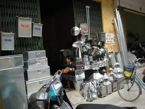 Bản ghi chữ viết:。- Trên các hòm thư bằng kim loại có dòng chữ "THƯ BÁO" màu đỏ và xanh lam.。- Trên bức tường màu vàng có một tấm biển ghi: "BÁN THIẾC - CHÌ - KẼM NHÔM & TÔN". Bên dưới có các số điện thoại, trong đó có một số đọc được là "0976 055...".。Mô tả cảnh vật và hoạt động:。Đây là khung cảnh phía trước một cửa hàng chuyên bán các sản phẩm làm từ kim loại tấm (sắt tây, tôn) trên một con phố. Cửa hàng có cửa xếp bằng sắt màu xanh lá cây, hiện đang mở. Một người phụ nữ đang ngồi trên một chiếc ghế đẩu nhỏ màu đỏ ngay lối ra vào, có vẻ như đang trông coi cửa hàng.。Các sản phẩm được trưng bày tràn ra cả vỉa hè, bao gồm:。- Các hòm thư bằng tôn có chữ "THƯ BÁO" được treo trên cửa.。- Nhiều loại rương, hòm tôn với các kích cỡ khác nhau được xếp chồng lên nhau.。- Các loại chụp thông gió mái nhà, ống khói, xô, phễu, và các vật dụng nhà bếp khác bằng kim loại sáng bóng.。- Các ống kim loại dài được dựng vào tường.。Phía trước có một chiếc xe máy đang đỗ, và bên phải là một chiếc xe đạp màu xanh lam có giỏ xe. Toàn bộ khung cảnh thể hiện hoạt động kinh doanh của một cửa hàng nhỏ, đặc trưng ở Việt Nam.。（日本語訳）。文字の記録：。- 金属製の郵便受けには、赤色と青色で「THƯ BÁO」という文字が書かれている。- 黄色い壁には、「BÁN THIẾC - CHÌ - KẼM NHÔM & TÔN」（錫・鉛・亜鉛・アルミニウム・トタン販売）と書かれた看板がある。その下には電話番号がいくつかあり、そのうちの一つは「0976 055...」と読み取れる。風景と活動の描写：。これは、ある通りにある、板金（ブリキ、トタン）製品を専門に販売する店の前の光景である。店には緑色の鉄製シャッタードアがあり、現在は開いている。一人の女性が、出入り口のすぐそばにある小さな赤いスツールに座っており、店番をしているようだ。商品は歩道にまであふれ出るように陳列されており、以下のようなものが含まれる：。- ドアに掛けられた、「THƯ BÁO」という文字が入ったトタン製の郵便受け。- 様々なサイズのトタン製の箱やケースが積み重ねられている。- 屋根の換気フード、煙突、バケツ、漏斗、その他の光沢のある金属製の台所用品。- 壁に立てかけられた長い金属パイプ。手前にはバイクが一台駐車しており、右側にはカゴ付きの青い自転車がある。全体の光景は、ベトナム特有の小さな店の営業活動を表している。