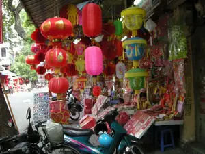 Trên một cây cột hoặc hộp các-tông ở bên phải có ghi các số điện thoại: "0913023234 096..." và một số chữ khác nhưng không thể đọc rõ. Có một tấm biển hiệu phía trên cửa hàng nhưng chữ bị mờ và không thể đọc được.。Đây là khung cảnh một cửa hàng ven đường ở Hà Nội, có khả năng nằm trên một khu phố chuyên bán đồ trang trí lễ hội như phố Hàng Mã. Cửa hàng đang bày bán rất nhiều loại đèn lồng và đồ chơi để chuẩn bị cho Tết Trung Thu. Vô số đèn lồng với đủ màu sắc và hình dạng được treo dày đặc trước cửa hàng, tạo nên một cảnh tượng rực rỡ. Có những chiếc đèn lồng giấy màu đỏ truyền thống hình tròn và hình trụ, một số có trang trí hoa văn màu vàng hoặc chữ Hán (ví dụ như chữ 'Tài'). Ngoài ra còn có đèn lồng màu hồng, đèn lồng giấy xếp hình tổ ong, và các loại đèn lồng nhiều tầng màu vàng, xanh lam, xanh lá cây, thường dành cho trẻ em. Bên trong và trên bàn bày bán còn có nhiều món đồ trang trí khác được bọc trong túi ni lông và các hộp quà màu đỏ. Một người phụ nữ đang ngồi bên trong cửa hàng trông coi hàng hóa. Phía trước có một vài chiếc xe máy đang đỗ. Ở phía xa trên con đường, có thể thấy nhiều người và xe cộ đang qua lại, cho thấy một khung cảnh đường phố nhộn nhịp.。（日本語訳）。店の上には看板がありますが、文字がぼやけていて読むことができません。これはハノイの道端にある店の光景で、おそらくハンマー通り（Hàng Mã）のような祭りの装飾品を専門に売る通りに位置しているのでしょう。この店は中秋節（Tết Trung Thu）の準備のために、たくさんの種類の提灯やおもちゃを並べて売っています。あらゆる色や形の無数の提灯が店の前にびっしりと吊るされ、きらびやかな光景を作り出しています。伝統的な赤色の丸い形や円筒形の紙提灯があり、その中には黄色の模様や漢字（例えば「財」という字）で装飾されているものもあります。その他にも、ピンク色の提灯、蜂の巣のような形の蛇腹提灯、そして黄色、青、緑の多層式の提灯があり、これらは通常子供向けです。店内や陳列台の上には、ビニール袋に包まれた他の多くの装飾品や赤いギフトボックスもあります。一人の女性が店の中で座って商品を番しています。前方には数台のバイクが停まっています。遠くの路上では、多くの人々や車が行き交っているのが見え、にぎやかな街の様子がうかがえます。