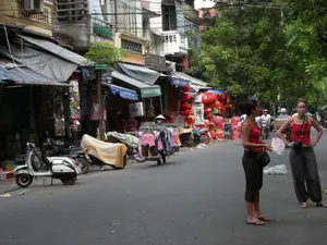 Bối cảnh là một con phố ở Hà Nội, với các tòa nhà ống cũ và cây xanh hai bên đường. Trên một cột có biển báo đường phố màu xanh lá cây ghi chữ 'PHỐ HÀNG MÃ'. Các cửa hàng ven đường có mái hiên, bày bán nhiều loại hàng hóa. Một cửa hàng có biển hiệu màu xanh dương ghi 'THỜI TRANG'. Một cửa hàng khác bán rất nhiều đèn lồng màu đỏ, một số có hình trái tim. Một người bán hàng rong đội nón lá đang đẩy xe hàng bán quần áo. Trên vỉa hè có một chiếc xe tay ga cổ màu trắng đang đậu. Giữa đường có hai nữ du khách nước ngoài đang đứng, một người cầm một chiếc quạt giấy. Phía sau có nhiều người đang di chuyển bằng xe máy.。（日本語訳）。背景はハノイのある通りで、古いチューブハウスと通りの両側に緑の木々があります。一本の柱には、「PHỐ HÀNG MÃ」と書かれた緑色の通りの標識があります。道端の店には日よけがあり、多くの種類の商品を陳列販売しています。ある店には「THỜI TRANG」と書かれた青い看板があります。別の店ではたくさんの赤い提灯を売っており、そのうちのいくつかはハート形です。ノンラーをかぶった一人の露天商が、服を売る屋台のカートを押しています。歩道には一台の白色の古いスクーターが停まっています。道の真ん中には二人の外国人女性観光客が立っており、一人は紙の扇子を持っています。後ろには多くの人々がバイクで移動しています。