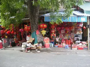 Đây là một khung cảnh trên một con phố ở Hà Nội, có vẻ như đang trong dịp chuẩn bị cho Tết Trung Thu. Ven đường, dưới một tán cây lớn, có các cửa hàng và người bán hàng rong. Các cửa hàng bày bán rất nhiều mặt hàng lễ hội, nổi bật là vô số đèn lồng màu đỏ với đủ loại hình dáng và kích cỡ được treo dày đặc. Ngoài đèn lồng tròn truyền thống, còn có thể thấy các loại đèn và đồ chơi Trung thu khác. Một cửa hàng có mái hiên sọc xanh trắng. Một người phụ nữ đang ngồi trên ghế đẩu màu xanh để bán hàng. Dưới gốc cây, một người bán hàng khác đội nón lá đang ngồi trên vỉa hè, bày hàng hóa trong những chiếc mẹt tre. Trên một số món đồ trang trí có thể thấy các chữ Hán như '囍' (Song Hỷ). Không khí trên phố cho thấy sự nhộn nhịp chuẩn bị cho một ngày lễ lớn.。（日本語訳）。これはハノイのある通りの光景で、中秋節（テト・チュントゥ）の準備期間中のようです。道端の、大きな木陰の下には、店や露天商がいます。店には多くのお祭りの品が並べられており、特に目立つのは、様々な形や大きさの無数の赤い提灯がびっしりと吊るされていることです。伝統的な丸い提灯の他に、他の中秋節の灯りやおもちゃも見られます。ある店には青と白の縞模様の軒先テントがあります。一人の女性が青いスツールに座って商品を売っています。木の根元では、ノンラーをかぶった別の売り子が歩道に座り、竹製の平たいざる（メット）に商品を並べています。いくつかの装飾品には、「囍」（ソンヒー、双喜）のような漢字が見られます。通りの雰囲気は、大きなお祭りに向けての賑やかな準備の様子を示しています。