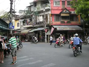 Bản ghi lại các thông tin từ hình ảnh:。1. Chữ viết được ghi lại:。- Trên mái hiên màu xanh lá cây bên trái: "VÍ DA - Ô DÙ CAO CẤP".。- Trên mái hiên của tòa nhà màu đỏ: "CỬA CUỐN CÁC LOẠI ĐẠI THẮNG". Một phần chữ khác không đọc rõ.。- Biển hiệu đèn neon màu xanh lá cây trên một tòa nhà: "DONG..." (phần còn lại không rõ).。- Có một biển báo giao thông màu xanh dương hình vuông chỉ dẫn lối cho người đi bộ qua đường.。- Có một biển báo giao thông hình tròn, viền đỏ, nền trắng (biển báo cấm).。2. Mô tả bối cảnh và hoạt động:。- Loại địa điểm: Đây là một góc phố hoặc ngã tư đông đúc tại một khu đô thị ở Việt Nam, có khả năng là Hà Nội. Các tòa nhà là kiểu nhà ống nhiều tầng, mặt tiền hẹp, san sát nhau. Tầng trệt của các tòa nhà được sử dụng làm cửa hàng kinh doanh.。- Hoạt động cụ thể: Bức ảnh ghi lại cảnh sinh hoạt và giao thông đường phố thường ngày. Rất nhiều người đang điều khiển xe máy (xe gắn máy) trên đường. Một số người đội mũ bảo hiểm. Có một nhóm người đi bộ, trong đó có vài người trông giống khách du lịch phương Tây, đang đứng ở góc đường. Giao thông trên đường khá tấp nập với nhiều xe máy di chuyển cùng lúc.。- Các vật thể trong ảnh: Các tòa nhà cao tầng với ban công, một trong số đó được sơn màu đỏ và có biểu tượng Âm Dương trên phần mái. Hệ thống dây điện chằng chịt là một đặc điểm nổi bật. Có nhiều cửa hàng với mái hiên, bao gồm một cửa hàng bán ví da, ô dù và một cửa hàng tên "ĐẠI THẮNG" bán cửa cuốn. Phương tiện chủ yếu là xe máy. Ngoài ra còn có cây xanh, cột điện và các biển báo giao thông.。（日本語訳）。画像からの情報の記録：。1. 記録された文字：。- 左側の緑色の軒先：「VÍ DA - Ô DÙ CAO CẤP」（革財布 - 高級傘）。- 赤い建物の軒先：「CỬA CUỐN CÁC LOẠI ĐẠI THẮNG」（ダイタン 各種巻き取りシャッター）。他の文字の一部ははっきりと読めない。- ある建物の緑色のネオンサイン：「DONG...」（残りの部分は不明）。- 歩行者横断用の通路を示す四角い青色の交通標識がある。- 赤い縁取りに白地の円形交通標識（禁止標識）がある。2. 背景と活動の説明：。- 場所の種類：これはベトナムの都市部、おそらくハノイの賑やかな街角または交差点である。建物は多層階のチューブハウス（間口が狭く奥行きが深い家）形式で、間口が狭く、密集している。建物の1階は店舗として使用されている。- 具体的な活動：写真は日常の生活風景と道路交通を記録している。非常に多くの人々が道路でバイク（原動機付自転車）を運転している。一部の人はヘルメットを着用している。道路の交通はかなり混雑しており、多くのバイクが同時に移動している。- 写真の中の物体：バルコニーのある高層ビル群。そのうちの一つは赤く塗られ、屋根部分に陰陽のシンボルがある。複雑に絡み合った電線システムが際立った特徴である。革財布や傘を売る店、そして「ĐẠI THẮNG」（ダイタン）という名前の巻き取りシャッターを売る店など、軒先のある多くの店がある。主な交通手段はバイクである。その他に、緑の木々、電柱、交通標識もある。