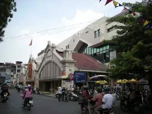 Bức ảnh chụp quang cảnh bên ngoài Chợ Đồng Xuân, một khu chợ lớn ở Hà Nội. Tòa nhà chợ có kiến trúc cổ với các cổng vòm lớn, phía sau là một khối nhà hiện đại hơn. Trên nóc tòa nhà có cắm lá cờ Việt Nam. Một biểu ngữ màu đỏ được treo trên mặt tiền chợ nhưng không đọc rõ chữ. Phía trước chợ là một con đường tấp nập người và xe cộ, chủ yếu là xe máy. Mọi người đang di chuyển trên đường, một số đội mũ bảo hiểm. Rất nhiều xe máy khác đang được dựng ở lề đường. Có một biển báo giao thông màu xanh lam ghi "CHỢ ĐỒNG XUÂN" cùng với các mũi tên chỉ đường và tên phố "HÀNG KHOAI". Bên cạnh là biển báo cấm dừng và đỗ xe. Dọc vỉa hè có những chiếc ô che nắng, cho thấy có hoạt động buôn bán nhỏ lẻ. Phía trên có treo một dãy cờ đuôi nheo nhiều màu sắc.。（日本語訳）。ハノイの大きな市場であるドン・スアン市場の外の光景を撮った写真です。市場の建物は大きなアーチ型の門を持つ古い建築様式で、その後ろにはより近代的な建物があります。建物の屋上にはベトナムの国旗が掲げられています。市場の正面には赤い横断幕が掲げられていますが、文字ははっきりと読めません。市場の前は、主にバイクを中心とした人や乗り物で賑わう道です。人々が道を移動しており、何人かはヘルメットをかぶっています。他の多くのバイクが道端に停められています。「CHỢ ĐỒNG XUÂN」と書かれた青い交通標識があり、方向を示す矢印と「HÀNG KHOAI」という通りの名前が記されています。その隣には、駐停車禁止の標識があります。歩道沿いには日よけの傘があり、小規模な商売が行われていることがわかります。上には色とりどりのペナントの列が飾られています。