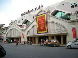 Bức ảnh chụp khung cảnh phía trước Chợ Đồng Xuân, một khu chợ lớn ở Hà Nội. Tòa nhà chợ có kiến trúc đặc trưng với các cổng vòm lớn. Trên nóc tòa nhà có dòng chữ lớn màu đỏ "CHỢ ĐỒNG XUÂN". Một tấm băng rôn lớn, sặc sỡ được treo ở mặt tiền tòa nhà với nội dung: "LỄ HỘI TRUNG THU PHỐ CỔ NĂM 2008". Trên đường phố phía trước chợ, có nhiều người và phương tiện giao thông như ô tô và xe máy đang hoạt động. Một số người đang đi bộ trên vỉa hè và dưới lòng đường. Có một chiếc taxi màu cam của hãng "TAXI CP" và một chiếc xe màu trắng của hãng "PHÚ ĐÔNG". Khung cảnh cho thấy hoạt động sinh hoạt thường ngày trên một con phố sầm uất, đồng thời có không khí chuẩn bị cho lễ hội.。（日本語訳）。ハノイの大きな市場であるドンスアン市場の前の風景を写した写真です。市場の建物は、大きなアーチ型の門が特徴的な建築様式です。建物の屋根には「CHỢ ĐỒNG XUÂN」という大きな赤い文字があります。建物の正面には「LỄ HỘI TRUNG THU PHỐ CỔ NĂM 2008」（2008年旧市街中秋節祭り）という内容の、大きくてカラフルな横断幕が掲げられています。市場の前の通りには、多くの人々や、自動車やバイクなどの交通手段が活動しています。歩道や車道を歩いている人もいます。「TAXI CP」社のオレンジ色のタクシーと、「PHÚ ĐÔNG」社の白い車があります。この風景は、賑やかな通りでの日常の営みを示すと同時に、祭りの準備の雰囲気も感じさせます。