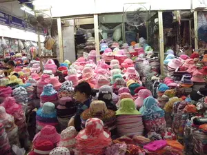 Bối cảnh là một gian hàng bên trong một khu chợ có mái che, có thể là ở Hà Nội. Gian hàng này chuyên bán các loại mũ nón. Vô số chiếc mũ với đủ loại màu sắc, kiểu dáng và chất liệu, chủ yếu là mũ vải, được xếp thành từng chồng cao. Các loại mũ bao gồm mũ rộng vành, mũ tai bèo và mũ cho trẻ em. Có một người phụ nữ đang ở giữa các chồng mũ, dường như đang xem hàng hoặc sắp xếp chúng. Phía trên cao có treo những chiếc quạt điện để làm mát không gian. Ở phía trên bên trái có một tấm biển hiệu màu xanh dương với chữ trắng nhưng không thể đọc rõ. Ở góc trên bên phải có một tấm biển nhỏ ghi số '09.'.。（日本語訳）。背景は、屋根付きの市場の中にある一軒の店で、おそらくハノイにあると思われます。この店は様々な種類の帽子を専門に販売しています。あらゆる種類の色、デザイン、素材の無数の帽子が、主に布製の帽子で、高く積み重ねられています。帽子の種類には、つば広の帽子、バケットハット、子供用の帽子が含まれます。帽子の山の間に一人の女性がおり、商品を見ているか、またはそれらを整理しているようです。頭上には空間を涼しくするために扇風機が吊るされています。左上には青地に白文字の看板がありますが、はっきりと読むことはできません。右上隅には「09.」という数字が書かれた小さな看板があります。