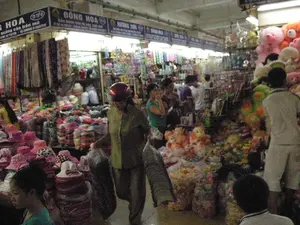 Chữ viết trên các biển hiệu:。- Biển hiệu bên trái: 'G HOA', 'quảng cáo trên mũ', 'ĐTHH: 04 7187238 - 04 CHỢ ĐỒNG XUÂN: 01689887988'.。- Biển hiệu ở giữa: 'ĐỒNG HÒA', 'Bán buôn - bán lẻ...'.。- Biển hiệu bên phải: 'HƯƠNG SƠN'.。Bối cảnh và hoạt động:。- Địa điểm: Đây là một khu chợ trong nhà, có thể là chợ Đồng Xuân ở Hà Nội dựa trên thông tin trên biển hiệu. Lối đi giữa các gian hàng khá hẹp và đông đúc người qua lại.。- Hoạt động: Khung cảnh cho thấy một phiên chợ tấp nập. Mọi người đang đi lại, xem hàng và mua sắm. Một người phụ nữ đội mũ bảo hiểm màu đỏ ở giữa đang xách hai chiếc túi lớn, cho thấy cô ấy có thể vừa mua một lượng lớn hàng hóa hoặc đang vận chuyển hàng.。- Vật thể: Các gian hàng được trưng bày san sát nhau, chất đầy hàng hóa. Gian hàng bên trái chuyên bán các loại mũ (nón) đủ màu sắc và kiểu dáng, được xếp thành từng chồng cao. Gian hàng bên phải bán đồ chơi, nổi bật là rất nhiều thú nhồi bông (gấu bông màu hồng, cam) với đủ kích cỡ. Nhiều người dân đang có mặt tại chợ, tạo nên một không khí nhộn nhịp.。（日本語訳）。看板の文字：。- 左の看板：「G HOA」、「帽子への広告」、「ĐTHH（電話）：04 7187238 - 04 ドンスアン市場：01689887988」。- 中央の看板：「ĐỒNG HÒA」、「卸売 - 小売...」。- 右の看板：「HƯƠNG SƠN」。背景と活動：。- 場所：これは屋内の市場エリアで、看板の情報からハノイのドンスアン市場である可能性があります。各店舗間の通路はかなり狭く、多くの人々が行き交っています。- 活動：活気のある市場の様子がうかがえます。人々は歩き回り、商品を眺め、買い物をしています。中央にいる赤いヘルメットをかぶった女性は、大きな袋を2つ持っており、大量の商品を購入したか、商品を運んでいる可能性があります。- 物体：各店舗は互いに密集して陳列されており、商品でいっぱいです。左側の店舗は、様々な色やデザインの帽子（ノン）を専門に販売しており、高く積み重ねられています。右側の店舗はおもちゃを販売しており、特に様々なサイズのぬいぐるみ（ピンクやオレンジのテディベア）が多数目立ちます。多くの人々が市場におり、にぎやかな雰囲気を醸し出しています。