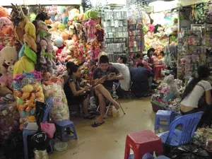 Bối cảnh là một gian hàng đông đúc, chật hẹp, có thể nằm trong một khu chợ hoặc trung tâm thương mại ở Hà Nội, chuyên bán đồ chơi. Không gian tràn ngập hàng hóa, chủ yếu là thú nhồi bông. Vô số thú nhồi bông với đủ kích cỡ, màu sắc và hình dạng (gấu, hươu, búp bê) được chất thành đống cao từ sàn lên gần trần nhà. Một số được đóng gói trong các túi ni lông lớn trong suốt. Ngoài ra, còn có các kệ trưng bày nhiều mặt hàng nhỏ khác được đóng gói trong bao bì nhựa. Nhiều người, có thể là người bán và khách hàng, đang có mặt trong cửa hàng. Họ ngồi trên những chiếc ghế nhựa thấp màu xanh và đỏ đặt trong lối đi nhỏ. Một số người đang xem điện thoại hoặc nghỉ ngơi, trong khi những người khác dường như đang lựa chọn hoặc sắp xếp hàng hóa. Không có văn bản hay biển hiệu nào có thể đọc được trong ảnh.。（日本語訳）。背景は、ハノイの市場かショッピングセンターにあると思われる、混雑して狭い、おもちゃを専門に売る店です。店内は商品で溢れており、主にぬいぐるみです。無数のぬいぐるみが、あらゆるサイズ、色、形（熊、鹿、人形）で、床から天井近くまで高く積み上げられています。いくつかは、大きな透明のビニール袋に詰められています。その他にも、プラスチックのパッケージに包装された他の多くの小物を陳列する棚があります。店員や客と思われる多くの人が店内にいます。彼らは狭い通路に置かれた青と赤の低いプラスチック製の椅子に座っています。写真には読み取れる文字や看板はありません。