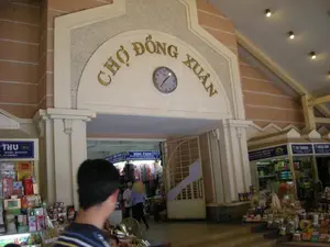 Bức ảnh chụp lối vào chính của Chợ Đồng Xuân, một khu chợ lớn có mái che ở Hà Nội. Phía trên cổng vào là một tấm biển lớn hình vòm với dòng chữ "CHỢ ĐỒNG XUÂN" bằng chữ nổi màu vàng. Trên biển hiệu có một chiếc đồng hồ đang chỉ khoảng 10 giờ 10 phút. Khung cảnh bên trong và xung quanh lối vào rất nhộn nhịp với nhiều người qua lại, trong đó có một người đang đội nón lá, cho thấy hoạt động mua bán, kinh doanh đang diễn ra. Hai bên lối vào là các quầy hàng san sát nhau. Ở bên trái, một quầy hàng có biển hiệu màu xanh ghi chữ "THU" và các thông tin "DTNR: 8233757", "19/06083.", bày bán nhiều sản phẩm đóng hộp. Ở bên phải, có các quầy hàng với biển hiệu mang tên "TƯ TRANG", "NGỌC ÁNH", và "BÌNH PHAN", trưng bày nhiều loại hàng hóa, chủ yếu là đồ khô và bánh kẹo được đóng gói. Bức ảnh ghi lại không khí mua sắm và kinh doanh tấp nập đặc trưng của khu chợ.。（日本語訳）。ハノイにある大きな屋根付き市場、ドンスアン市場の正面入口を撮影した写真です。入口ゲートの上には、「CHỢ ĐỒNG XUÂN」という文字が黄色の浮き彫りで書かれた大きなアーチ型の看板があります。看板の上には、10時10分頃を指している時計があります。入口の内外の光景は多くの人々が行き交い、非常に賑わっており、その中にはノンラーをかぶった人もいて、売買や商売が行われていることを示しています。入口の両側には、屋台がひしめき合っています。左側には、「THU」という文字と「DTNR: 8233757」、「19/06083.」という情報が書かれた青い看板のある屋台があり、多くの缶詰製品を陳列販売しています。右側には、「TƯ TRANG」、「NGỌC ÁNH」、「BÌNH PHAN」という名前の看板を掲げた屋台があり、主に乾物や包装された菓子類など、多くの種類の商品を陳列しています。この写真は、市場特有の賑やかな買い物と商売の雰囲気を捉えています。