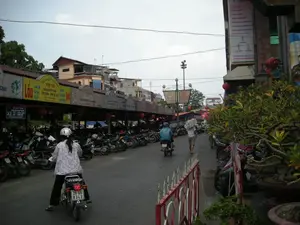 Bối cảnh là một con phố ở Hà Nội với nhiều xe máy và các quán ăn. Bên trái đường là một bãi giữ xe máy lớn, nơi hàng chục chiếc xe được xếp ngay ngắn thành hàng dài. Phía trên bãi giữ xe là một dãy các quán ăn liền kề có mái hiên chung, được trang trí bằng đèn lồng đỏ. Các biển hiệu quảng cáo món ăn rất nổi bật.。Các dòng chữ có thể đọc được trên các biển hiệu bao gồm:。- Biển trông giữ xe: "BẢNG GIÁ TRÔNG GIỮ XE ĐẠP VÀ XE MÁY", "XE ĐẠP: 1.000Đ/LƯỢT", "XE MÁY: 2.000Đ/LƯỢT".。- Biển hiệu các quán ăn: "...cháo", "LẨU", quán "PHƯƠNG LOAN" với các món "VỊT CỎ", "HẢI SẢN TƯƠI SỐNG", "CƠM RANG - PHỞ MỲ XÀO", "BÒ", "TƯƠI SỐNG", "HẤP". Các biển hiệu khác quảng cáo "Lẩu Các Món", "CƠM CHÁY".。- Ở phía xa có biển hiệu ghi "THỊT RỪNG".。- Trên tòa nhà bên phải có một tấm biển quảng cáo dọc màu hồng nhưng chữ bị mờ, không đọc được.。Hoạt động đang diễn ra trên đường phố bao gồm một người phụ nữ mặc áo hoa, đội mũ bảo hiểm đang lái xe máy; một người khác mặc áo mưa xanh cũng đang đi xe máy; và một người đàn ông đang đi bộ, vác một bao hàng lớn màu trắng trên vai. Phía bên phải vỉa hè có đặt nhiều chậu cây cảnh và một hàng rào sắt sơn màu đỏ và trắng.。（日本語訳）。背景はハノイのある通りで、たくさんのバイクと飲食店があります。道の左側には大きなバイク駐車場があり、そこでは数十台のバイクが長い列に整然と並べられています。駐車場の奥には、赤い提灯で飾られた、共通の軒先を持つ飲食店が軒を連ねています。料理を宣伝する看板がとても目立っています。看板に書かれている読み取れる文字は以下の通りです：。- 駐輪・駐車場の看板：「自転車・バイク駐輪駐車料金表」、「自転車：1,000ドン/回」、「バイク：2,000ドン/回」。- 飲食店の看板：「...cháo（お粥）」、「LẨU（鍋）」、「PHƯƠNG LOAN」という店には「VỊT CỎ（アヒル）」、「HẢI SẢN TƯƠI SỐNG（新鮮な海産物）」、「CƠM RANG - PHỞ MỲ XÀO（チャーハン - 焼きそば・フォー）」、「BÒ（牛）」、「TƯƠI SỐNG（新鮮）」、「HẤP（蒸し）」といった料理があります。他の看板には「Lẩu Các Món（各種鍋料理）」、「CƠM CHÁY（おこげ）」と宣伝されています。- 遠くには「THỊT RỪNG（ジビエ）」と書かれた看板があります。- 右側の建物にはピンク色の縦長の広告看板がありますが、文字がぼやけていて読めません。通りで行われている活動には、花柄の服を着てヘルメットをかぶった女性がバイクを運転している様子、青いレインコートを着た別の人がバイクに乗っている様子、そして白い大きな荷袋を肩に担いで歩いている男性の姿が含まれます。歩道の右側には、多くの観葉植物の鉢と、赤と白に塗られた鉄の柵が設置されています。